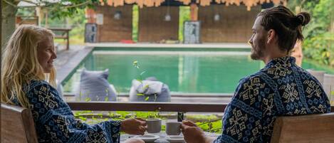 Two people sitting at a wooden table with a view of a swimming pool and a traditional building in the background.