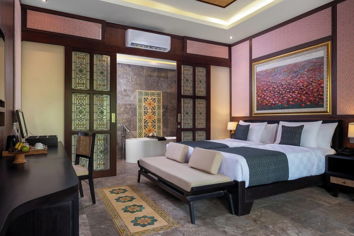 A hotel room with a bed, bedside tables, a desk, a chair, a sofa, a bathroom with a bathtub, and a large painting on the wall.
