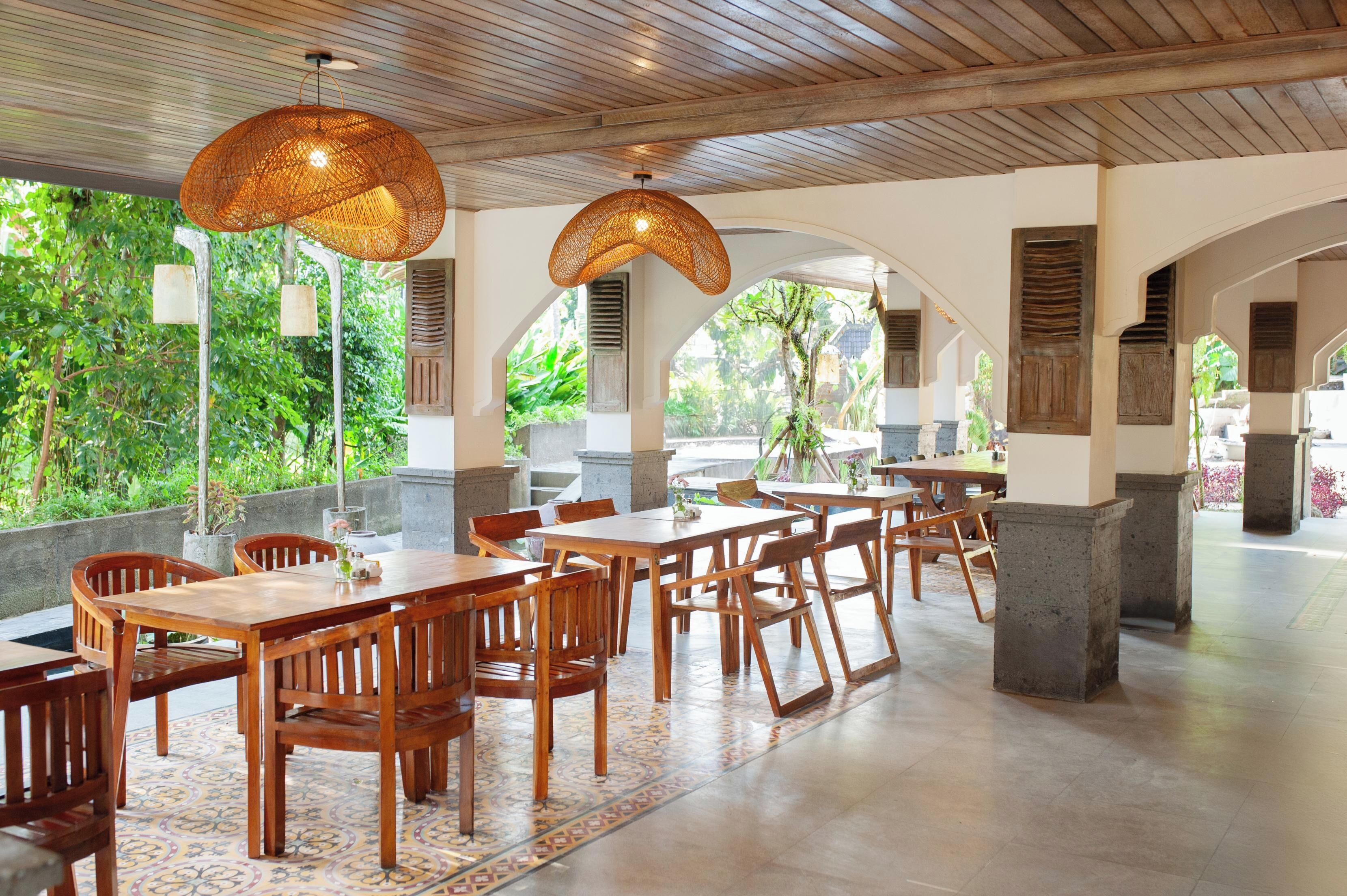 A spacious indoor dining area with wooden tables and chairs, large woven pendant lights, and a view of lush greenery outside.
