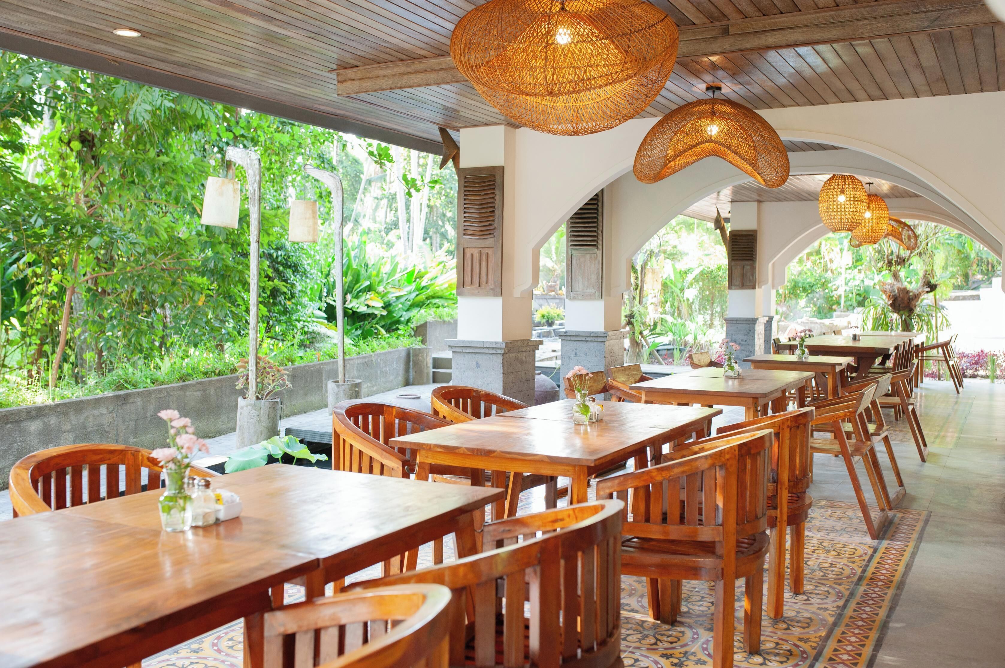 Un restaurant avec des tables et des chaises en bois, de grandes lampes suspendues en osier et une vue sur une végétation luxuriante.