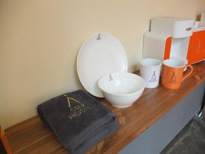 A hotel room's reception area with a coffee maker, a plate, a bowl, a mug, and a towel with the hotel's logo.