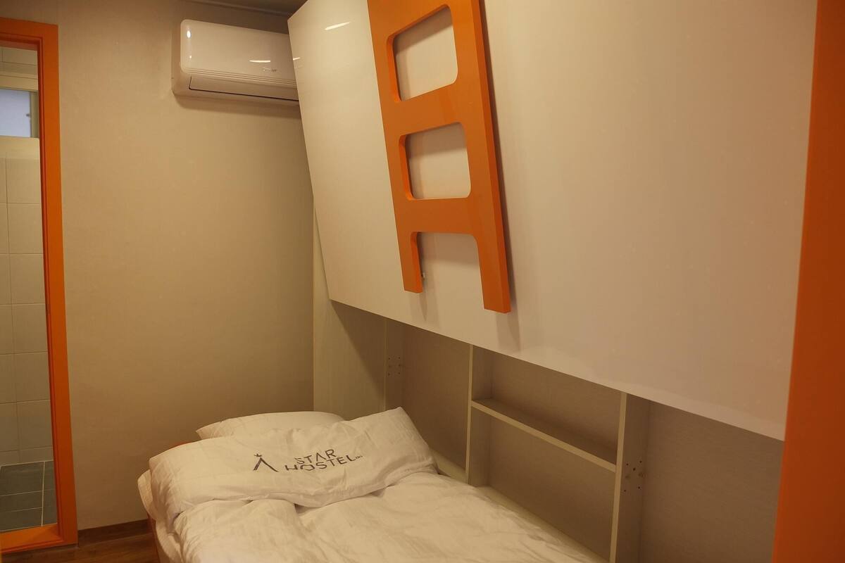Una cama pequeña y bien arreglada con colcha y almohadas blancas, una estantería de madera tipo escalera y un equipo de aire acondicionado blanco en la pared.