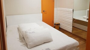 Une petite chambre avec un lit simple, une couette blanche, un oreiller blanc et une petite poubelle.
