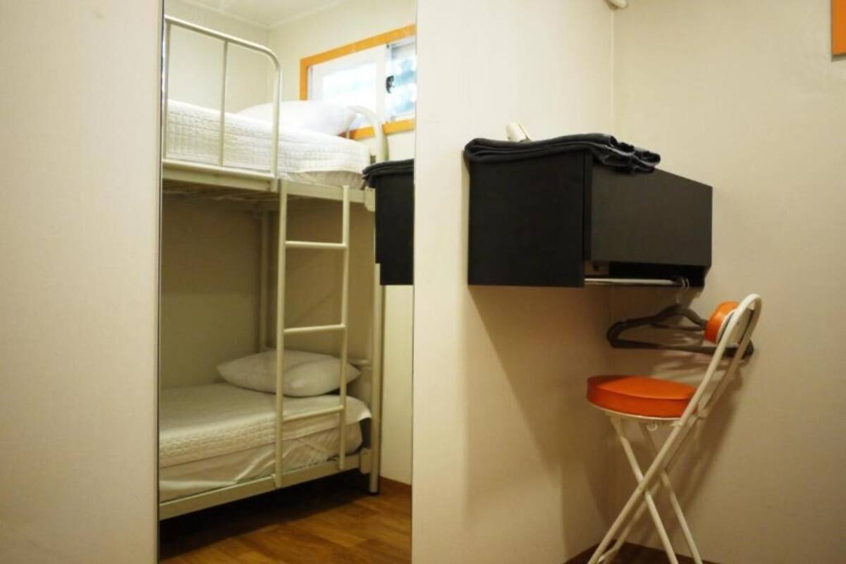 Una cama litera con escalera, una mesita con un objeto negro encima y una silla plegable.