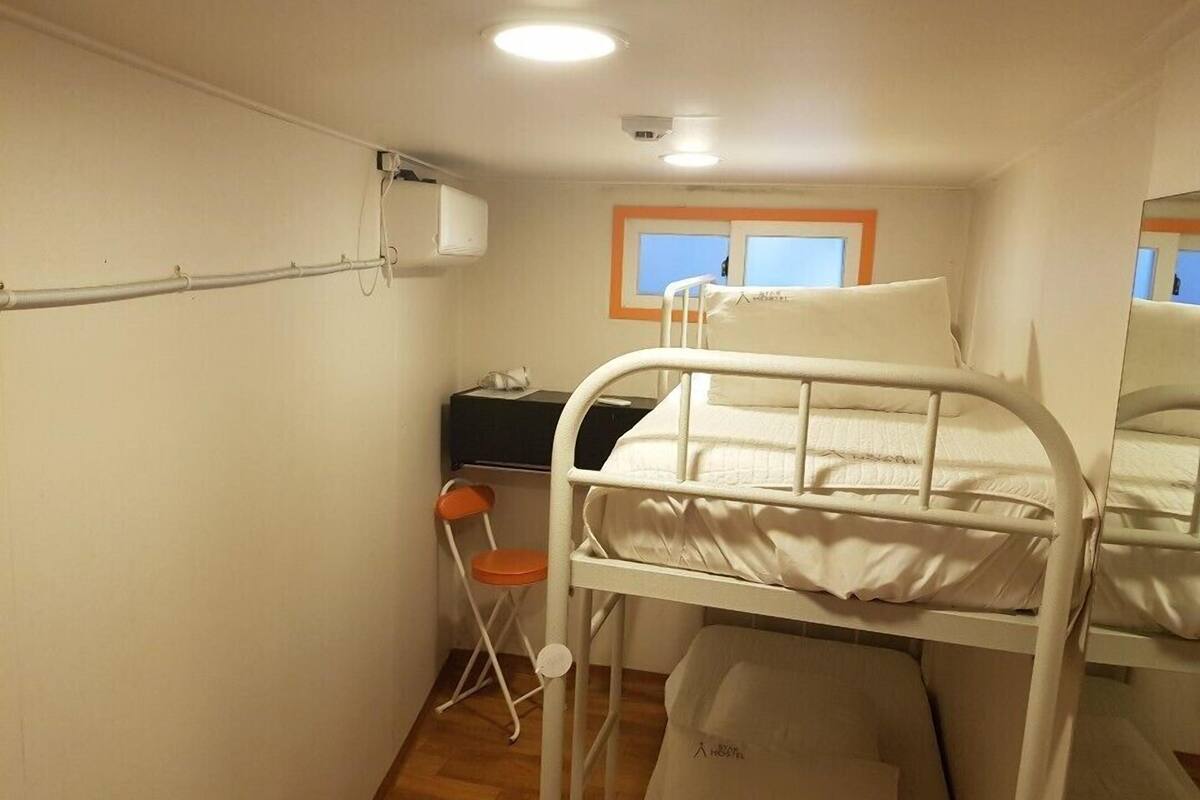 Habitación de hotel compacta con literas, un escritorio pequeño, una silla y una ventana con marcos naranja.