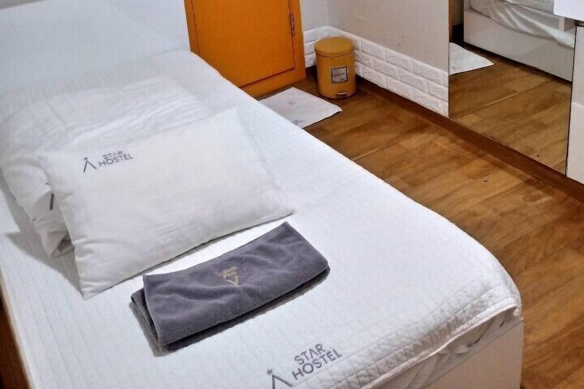 Habitación con una cama individual, suelo de madera, ropa de cama blanca, un cubo de basura pequeño y una puerta.
