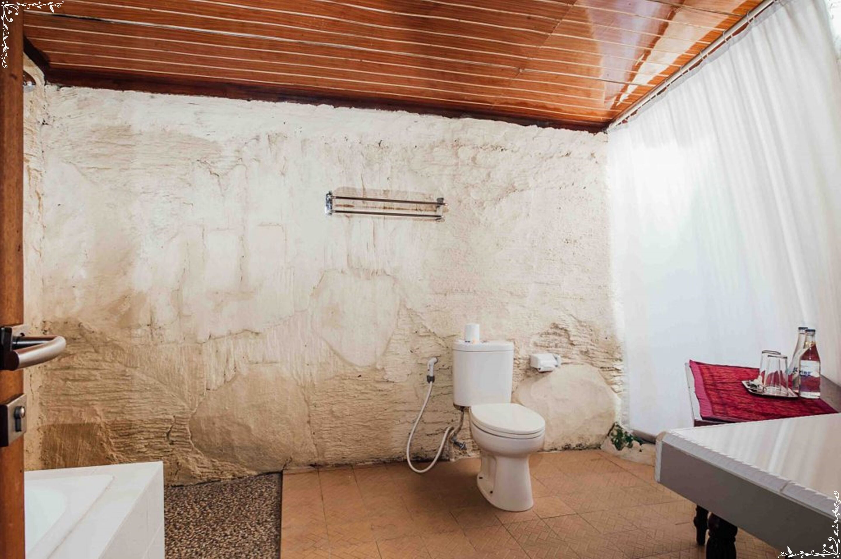 A small, rustic bathroom with a white toilet, a wooden ceiling, and a red towel on a small table.