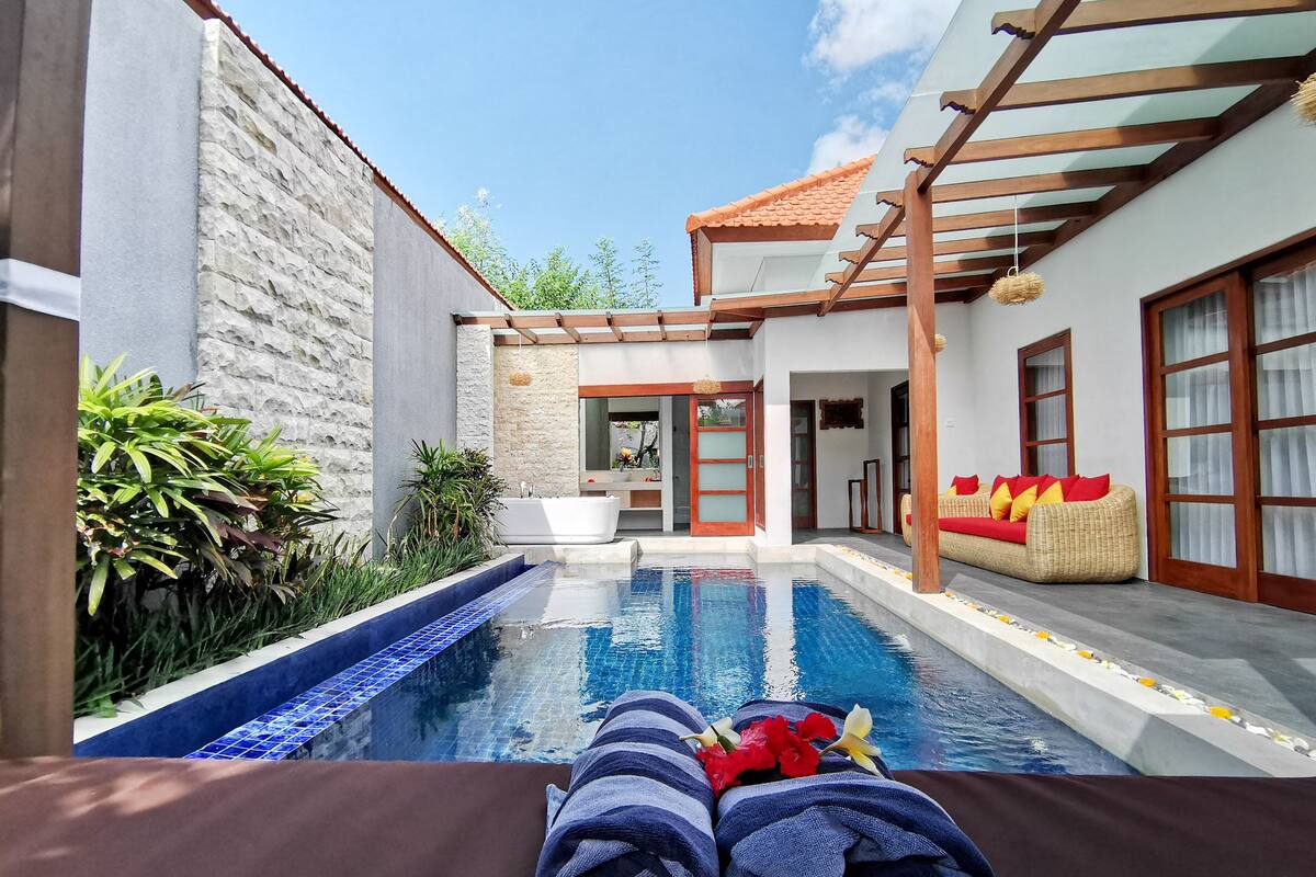A pool area with a stone wall, a wooden pergola, and a wicker sofa with red cushions.