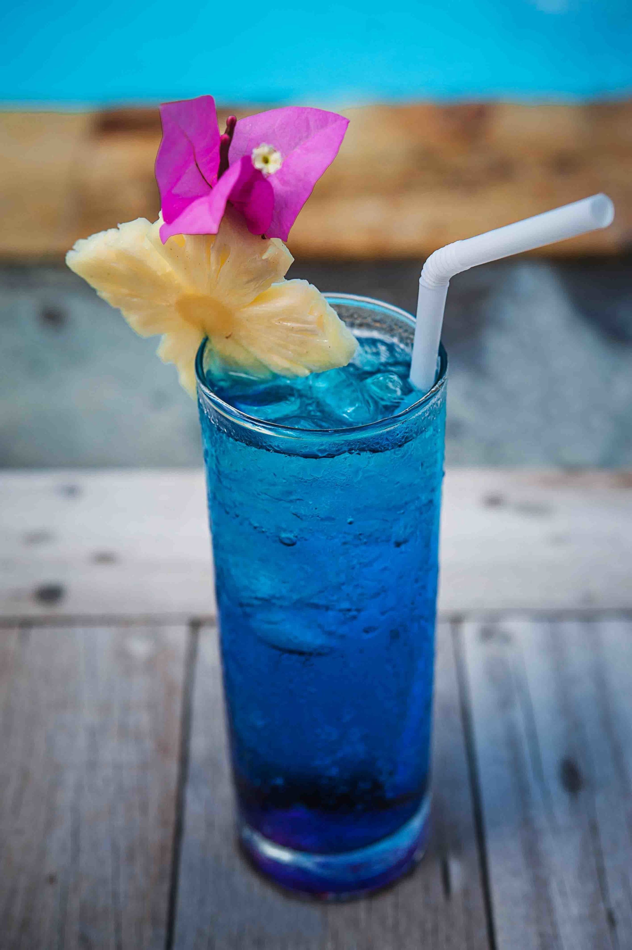 A tall glass with a blue beverage, a white straw, and a pineapple slice and pink flower garnish.