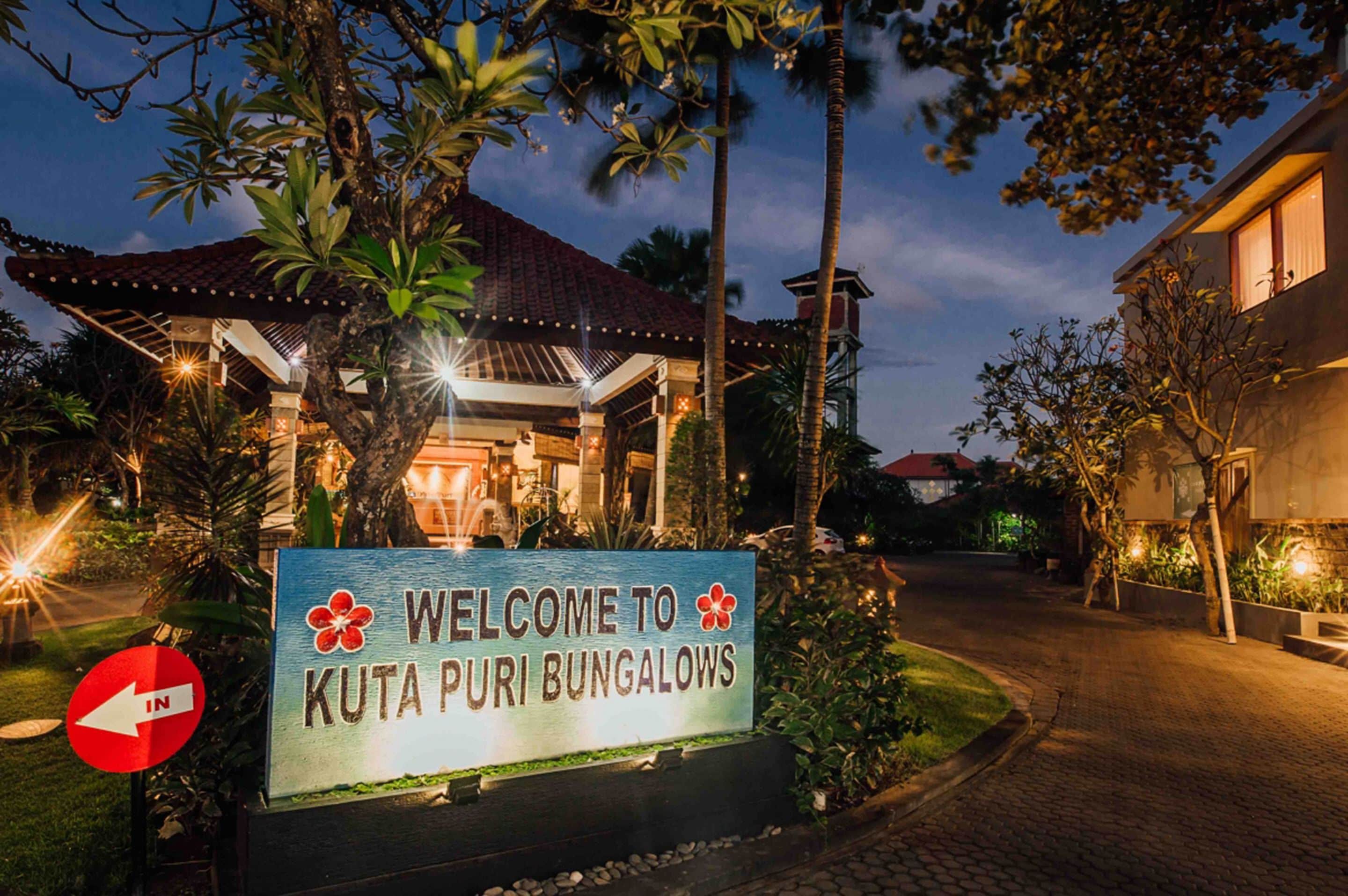 Ein Schild empfängt Besucher von Kuta Puri Bungalows, mit Blick auf das Gebäude und die umliegende Bepflanzung.