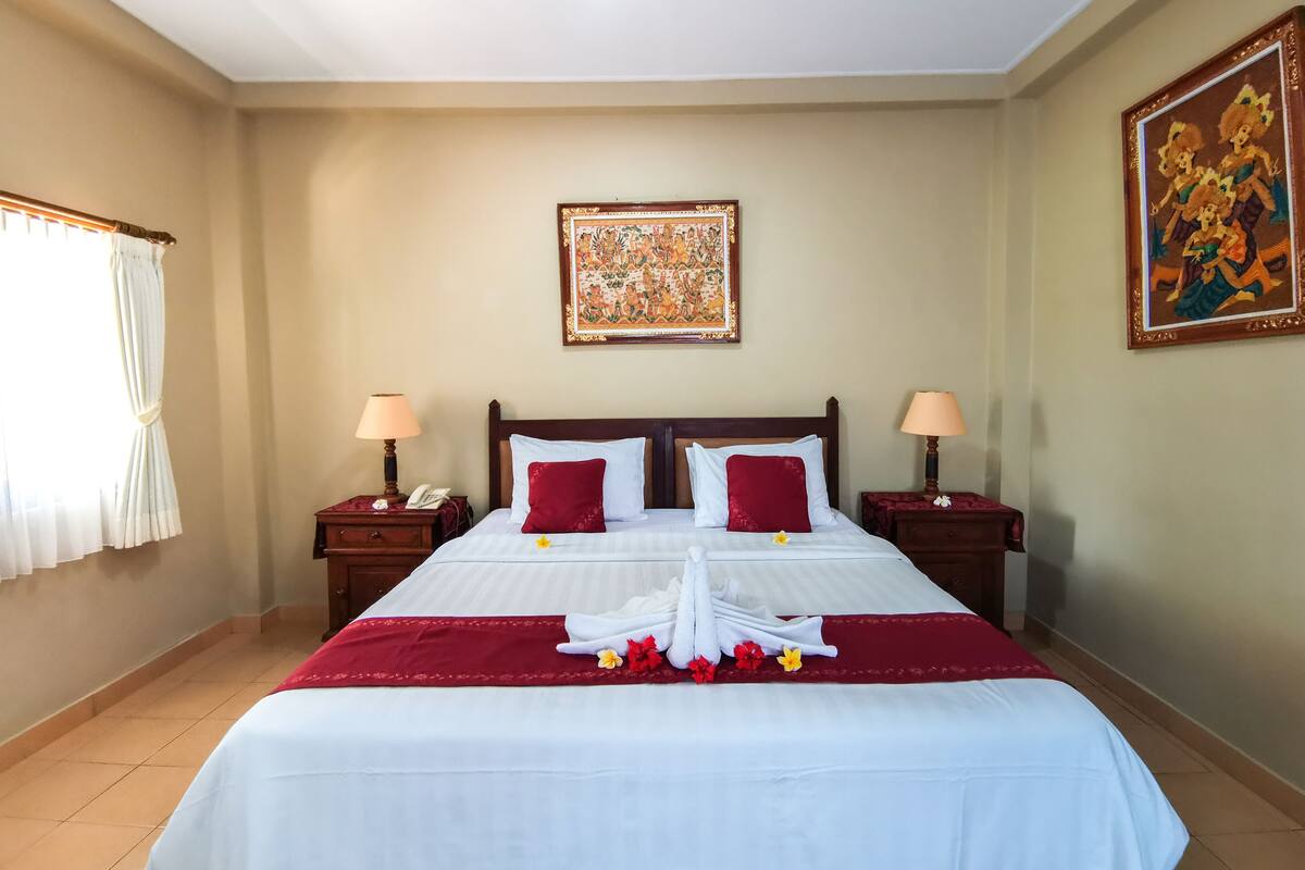 A hotel room with a large bed, two bedside tables, a nightstand, a window with curtains, and a wall with a framed painting.