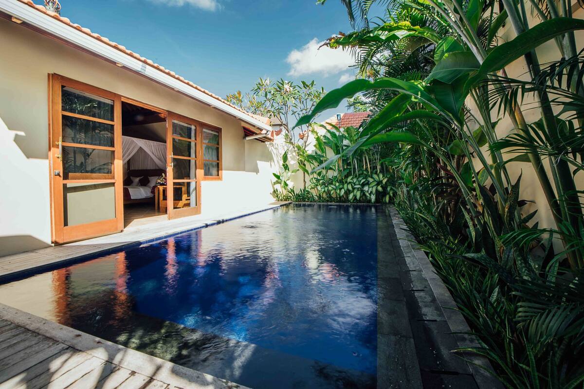 A pool area with a wooden deck, a white building with a sliding glass door, and lush tropical plants.