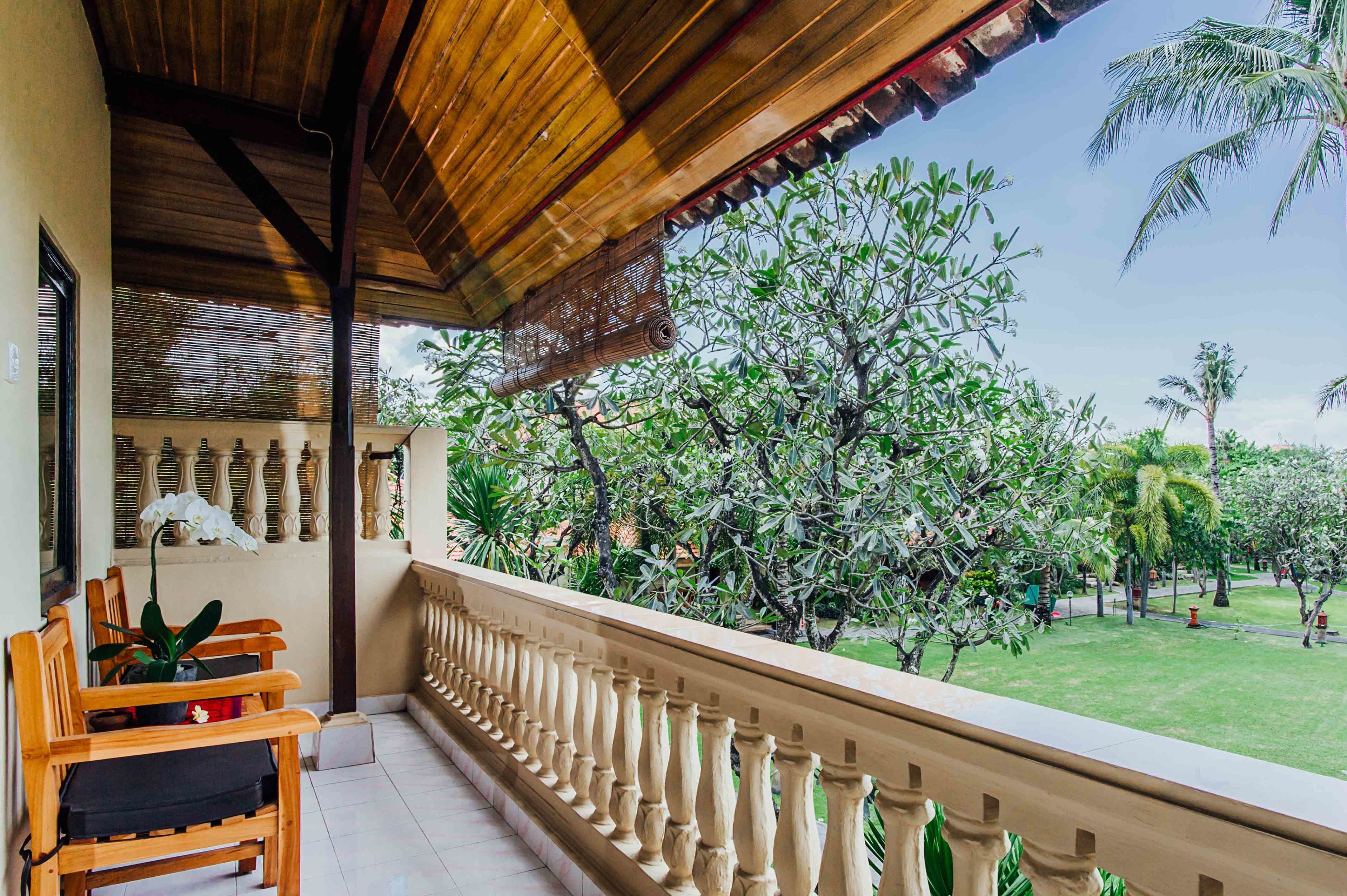 A balcony with wooden chairs and a potted plant, overlooking a well-maintained garden with various trees and a clear sky.