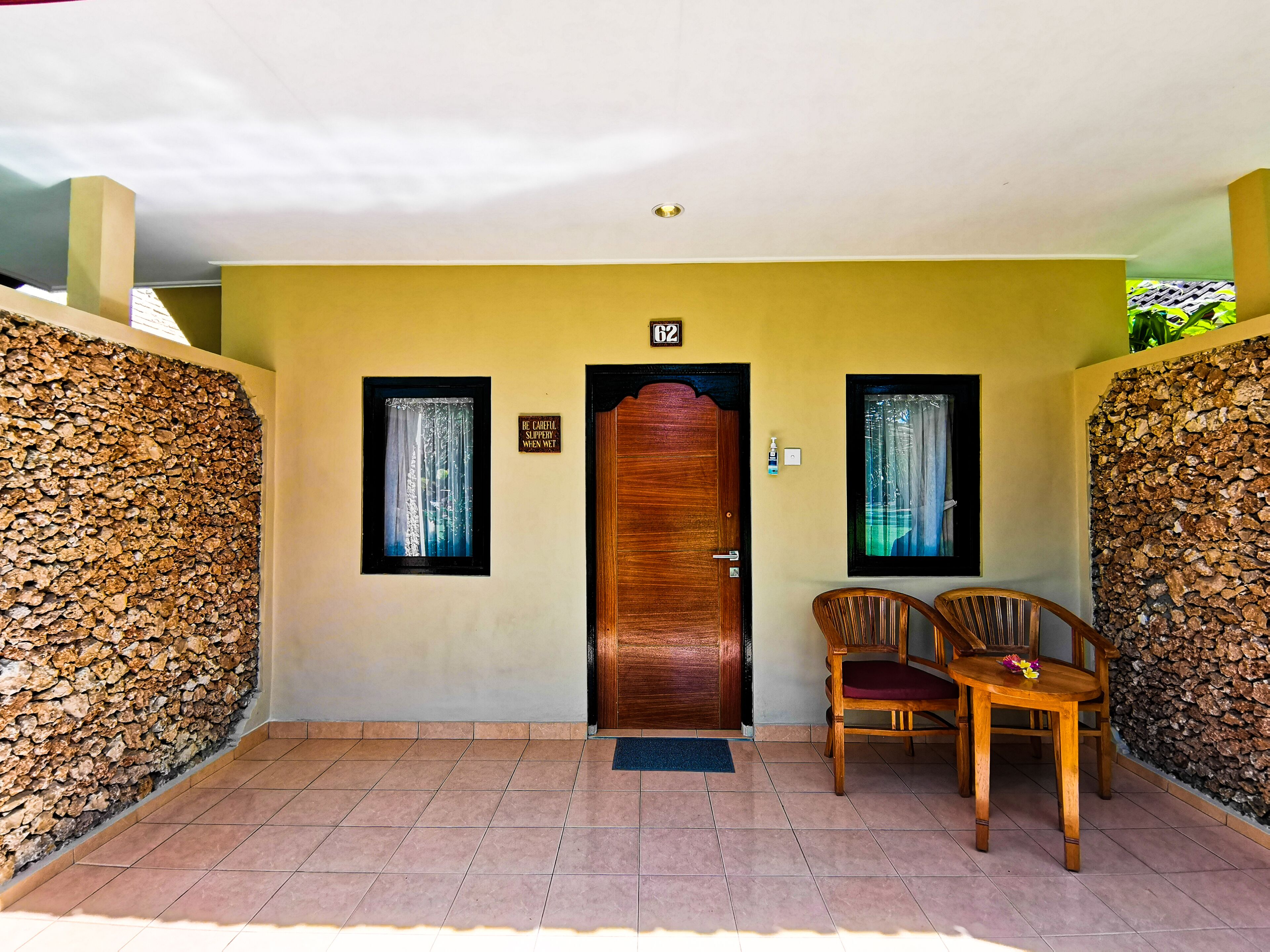 Fachada de hotel con puerta de madera, dos ventanas con cortinas, un cartel sobre la puerta y una pared de piedra a un lado.