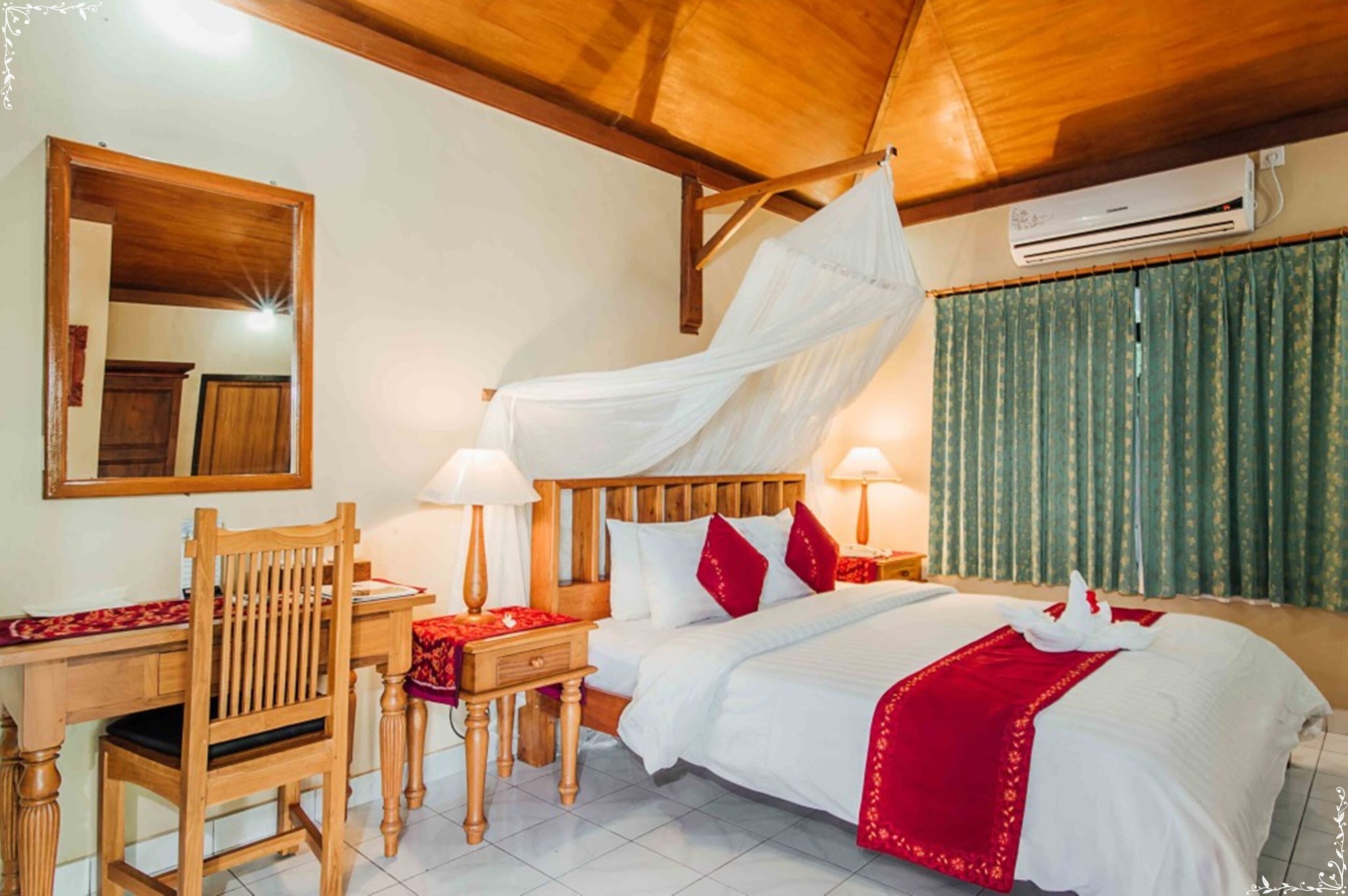 A hotel room with a wooden bed, white bedding, red pillows, a canopy bed, a desk with a chair, a mirror, and an air conditioning unit.