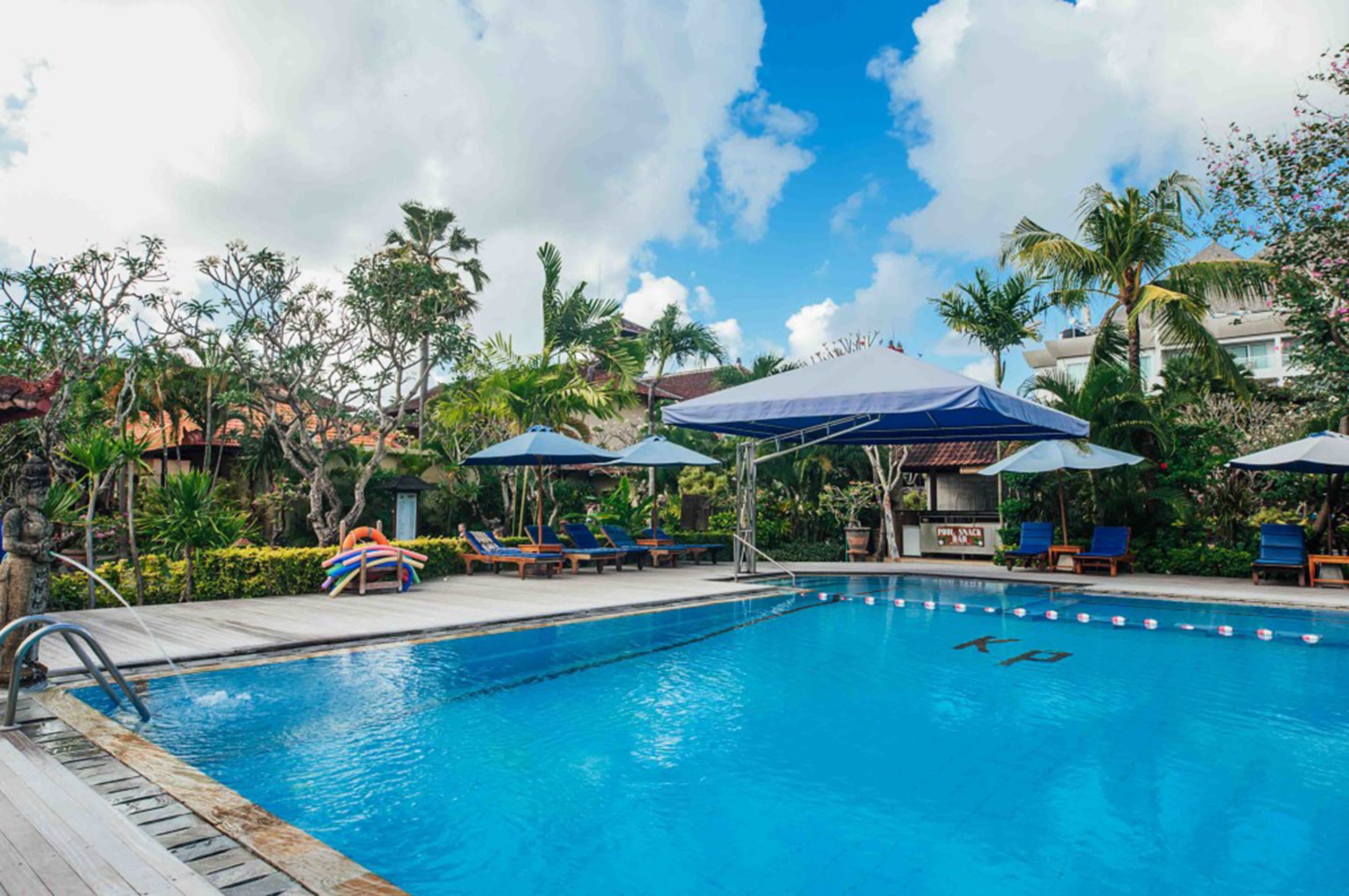 A swimming pool with lounge chairs and umbrellas, surrounded by tropical plants and a clear sky.