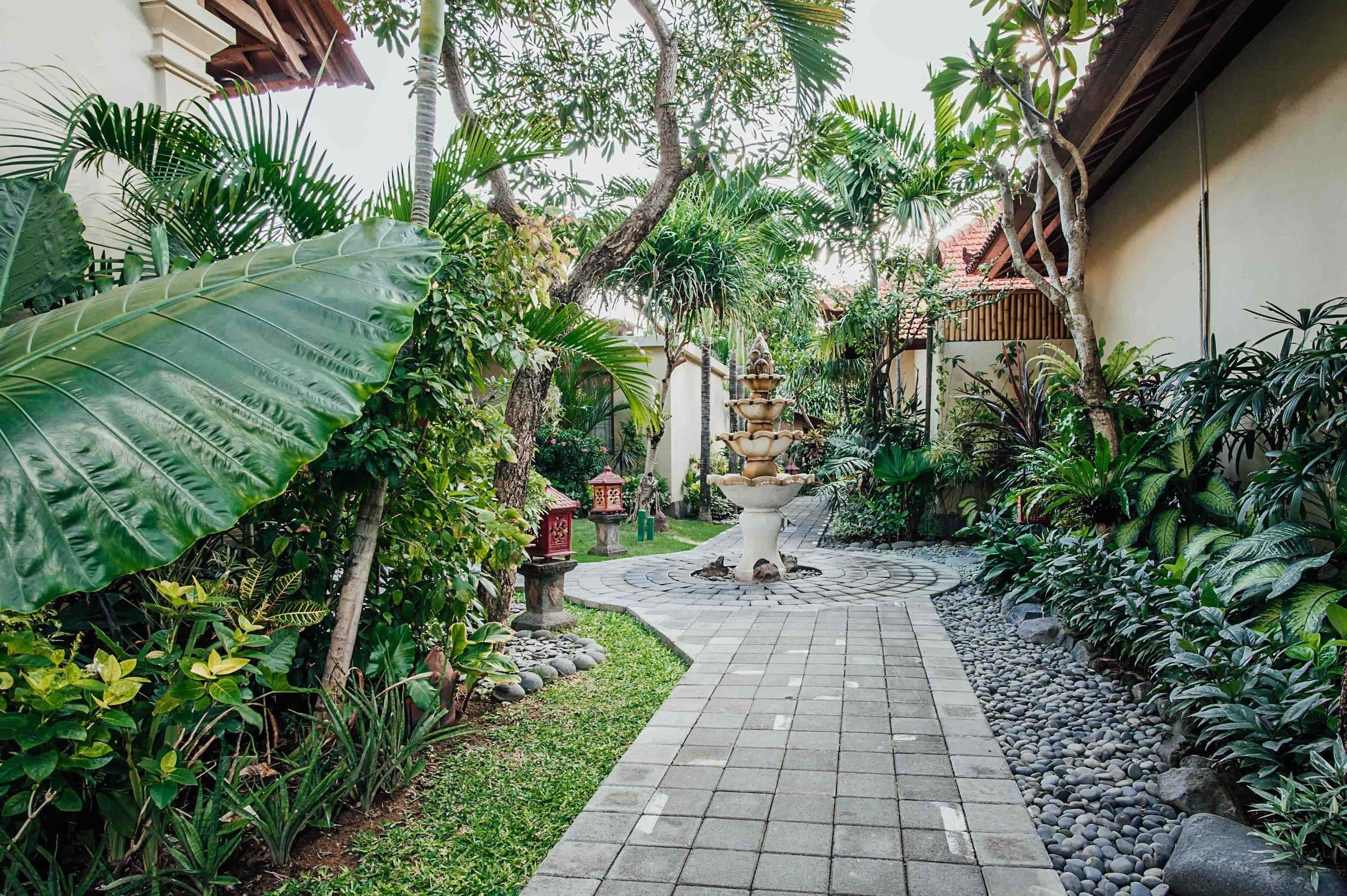 Un sendero de jardín con una fuente central, rodeado de exuberante vegetación tropical y un camino empedrado.