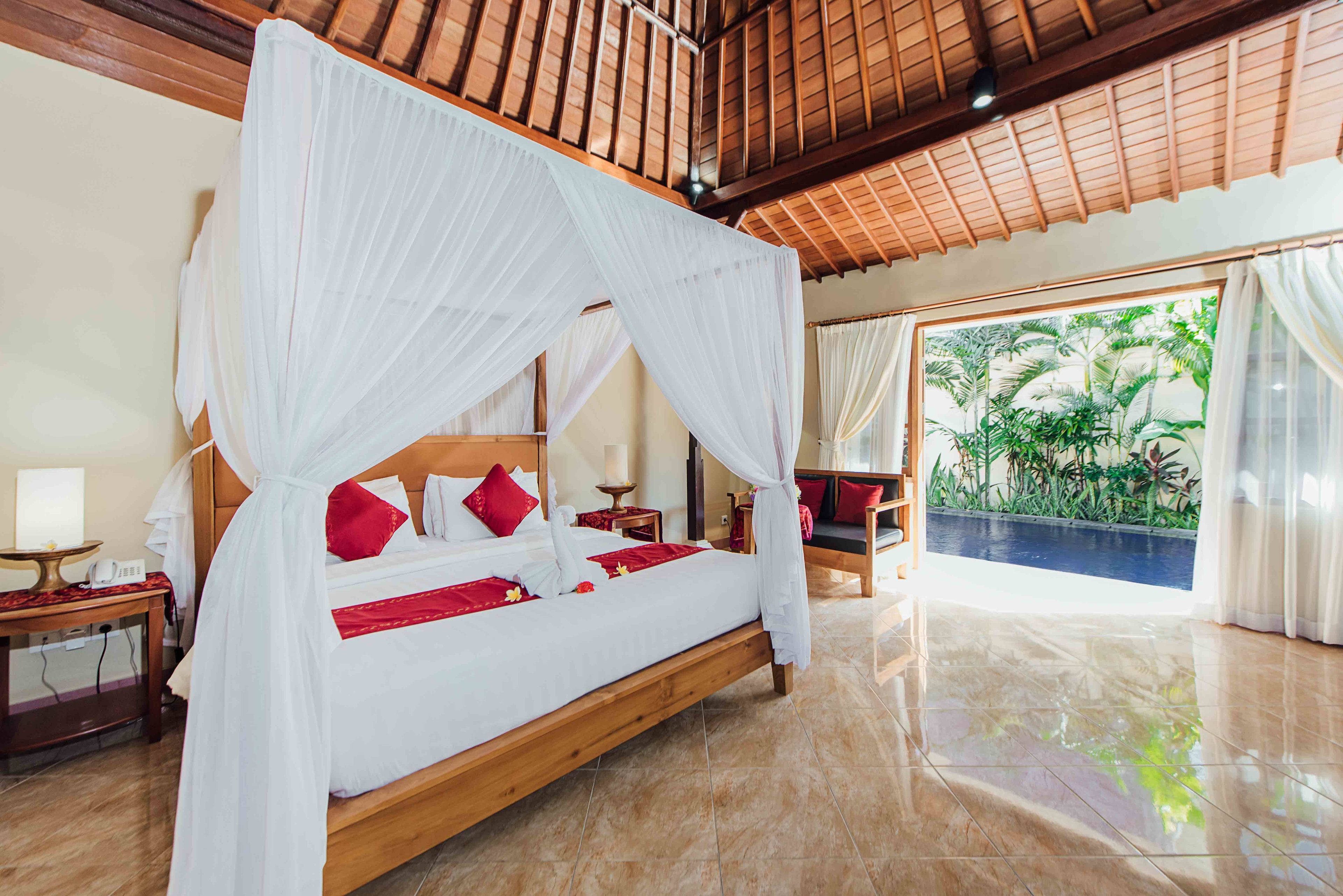 A spacious bedroom with a wooden bed, white canopy, red pillows, and a view of lush greenery through large windows.