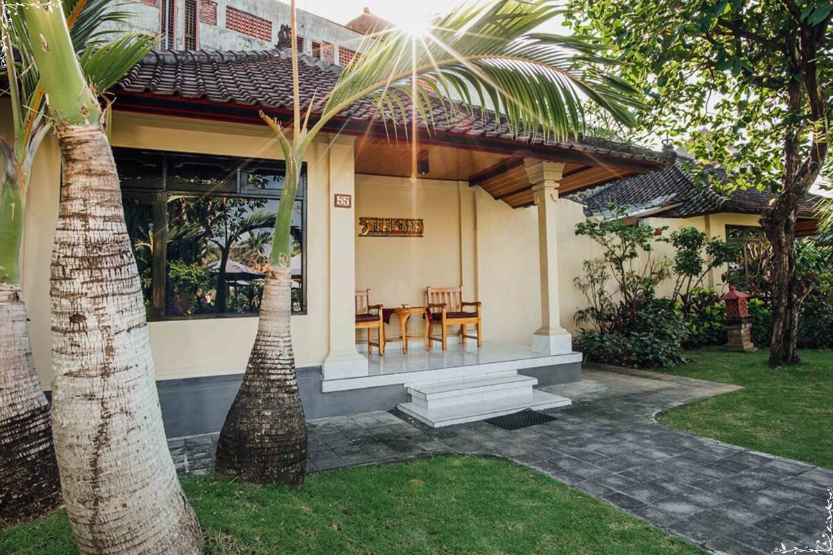 A traditional house with a tiled roof, a covered porch with wooden chairs, and a well-maintained garden with palm trees.