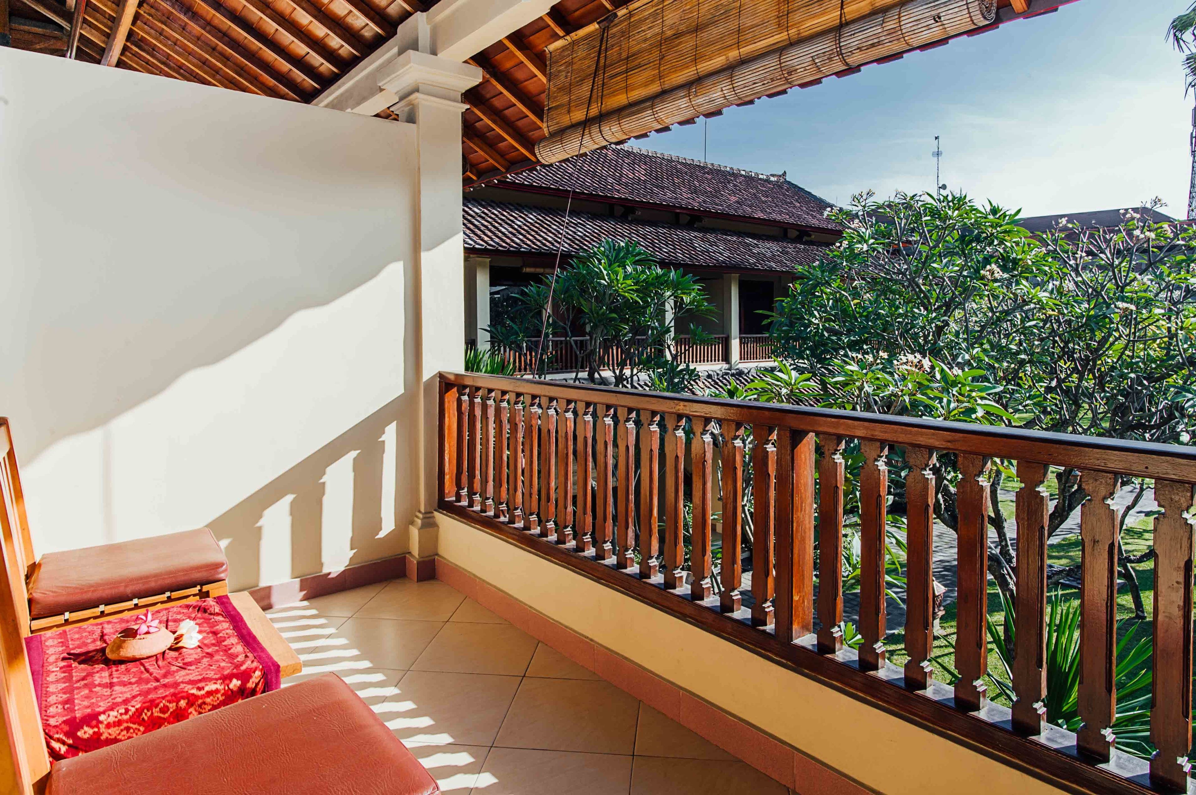 A balcony with a wooden railing, a red cushioned bench, and a view of a traditional building and greenery.