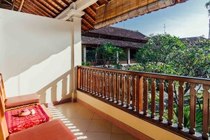 A balcony with a wooden railing, a red cushioned bench, and a view of a traditional building and greenery.