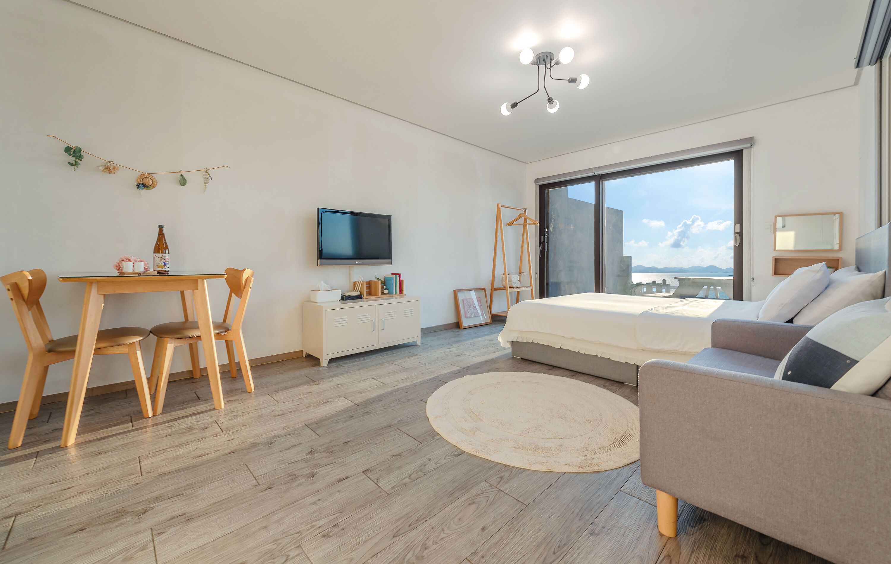 Quarto moderno com cama, sofá, televisão, escrivaninha com cadeiras e uma janela grande.