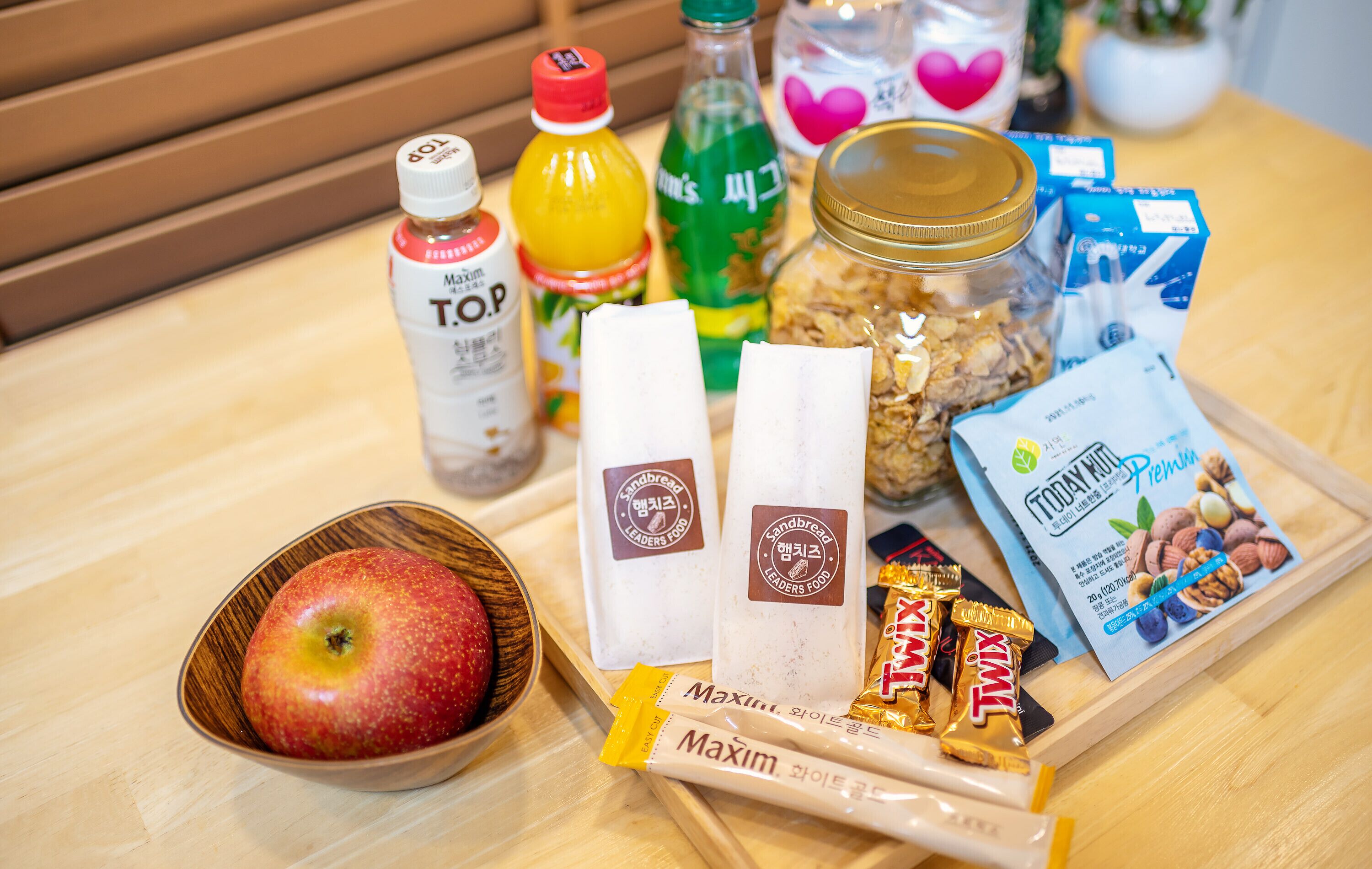 A wooden table with various food items including a bowl of apples, bottles of beverages, granola, and packaged snacks.