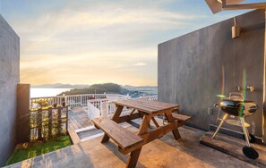 A rooftop patio with a wooden picnic table and a portable grill, overlooking a cityscape and hills. - Whale's Dream (Namhae)
