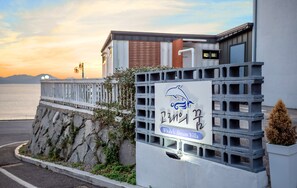 A sign for 'Whale's Dream Villa' with a dolphin logo, a stone wall, and a view of the sea at sunset. - Whale's Dream (Namhae)