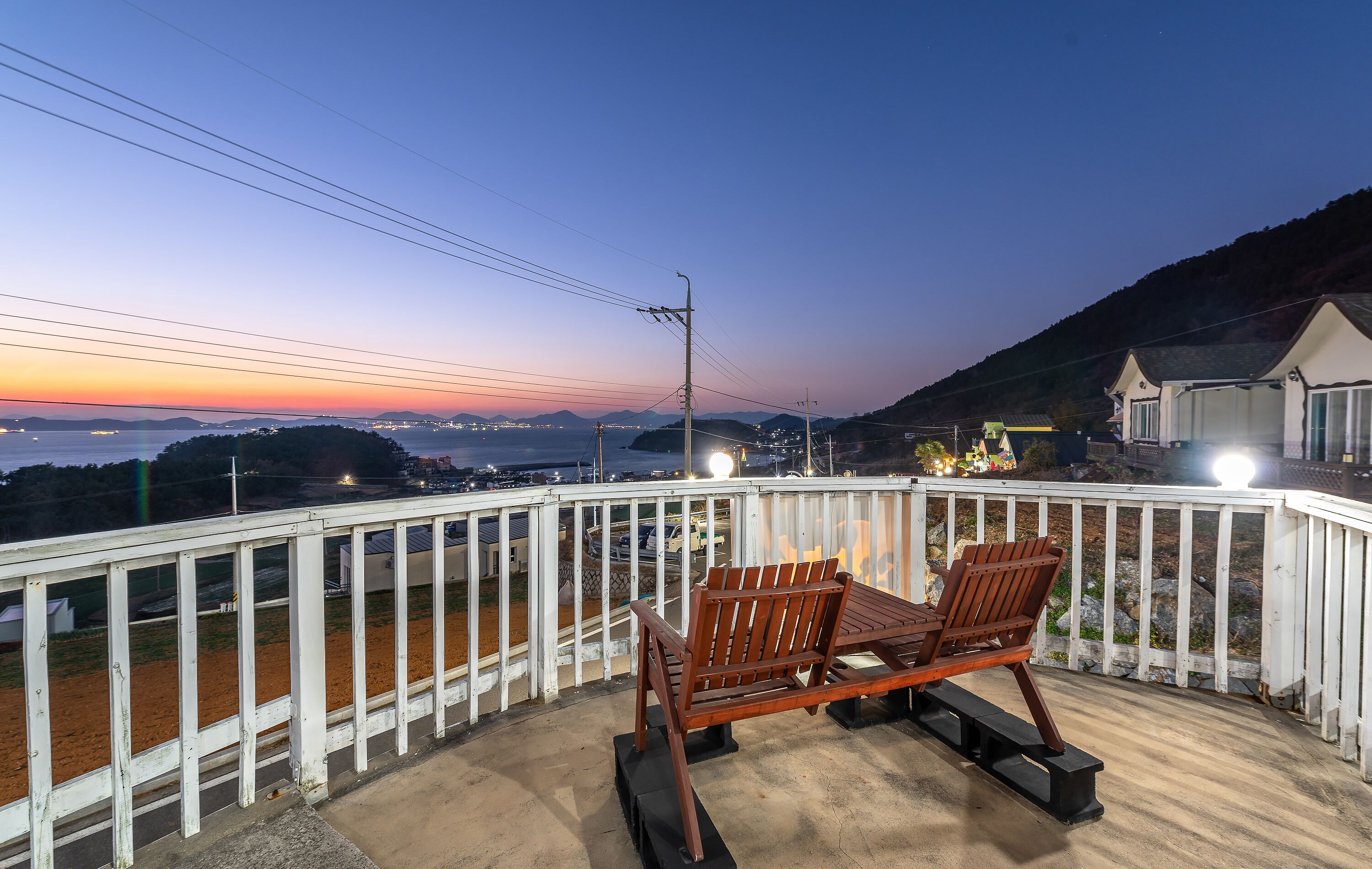 A balcony with a wooden table and chairs, overlooking a coastal area with distant mountains and a sunset.