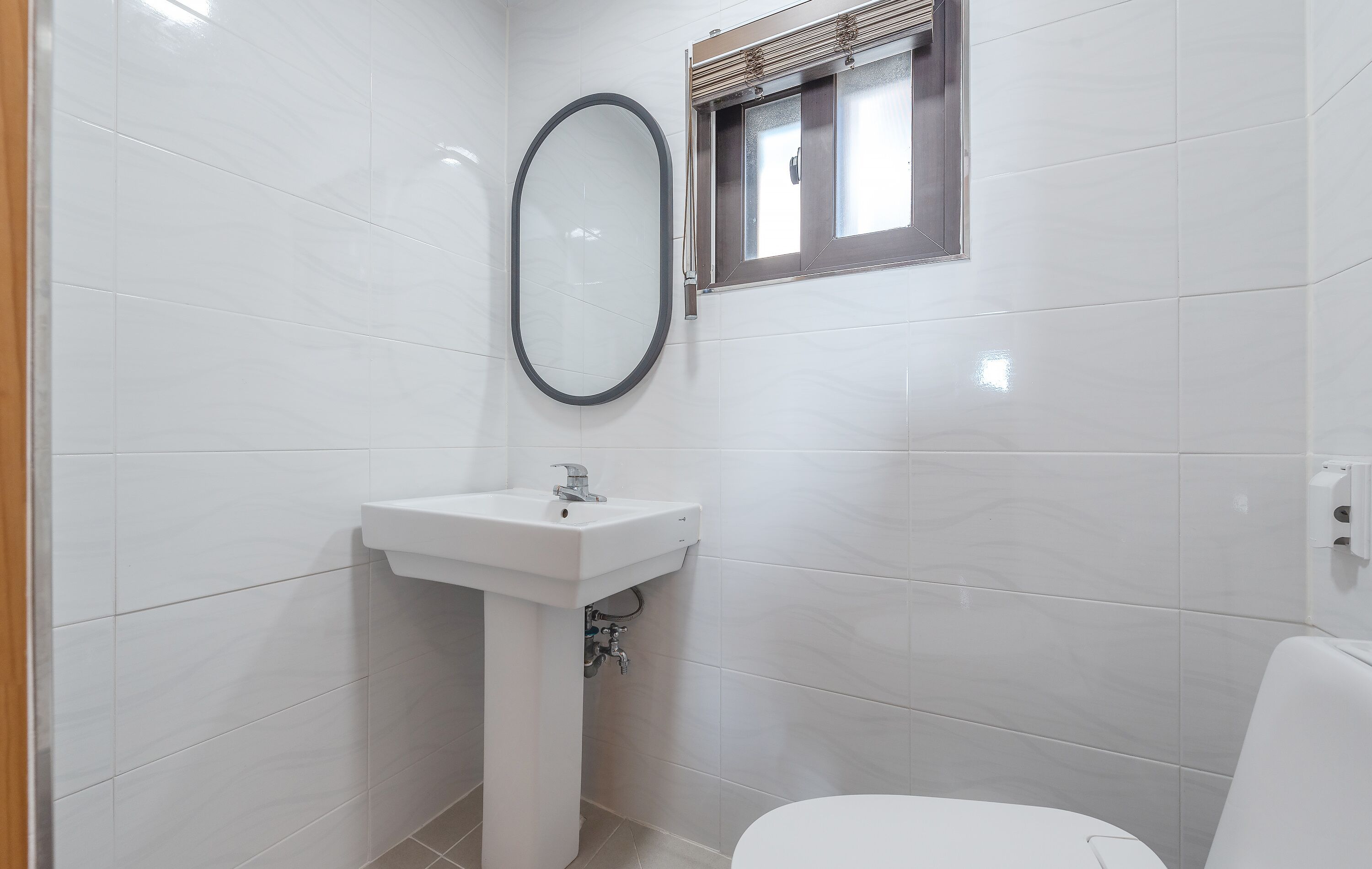 A small, modern bathroom with white tiles, a pedestal sink, a mirror, and a window with blinds.