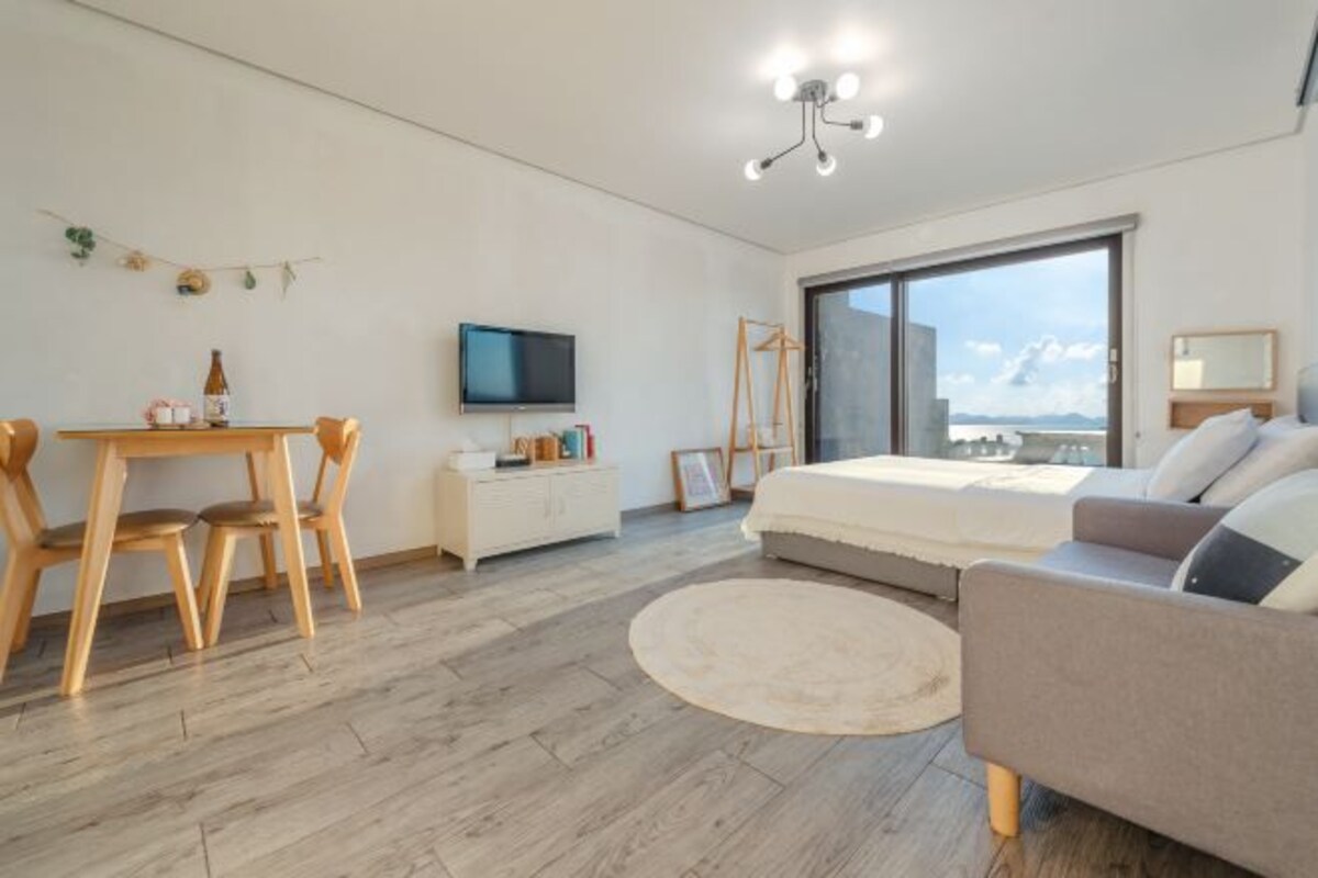 A modern bedroom with a bed, a TV, a desk with chairs, and a large window offering a view.