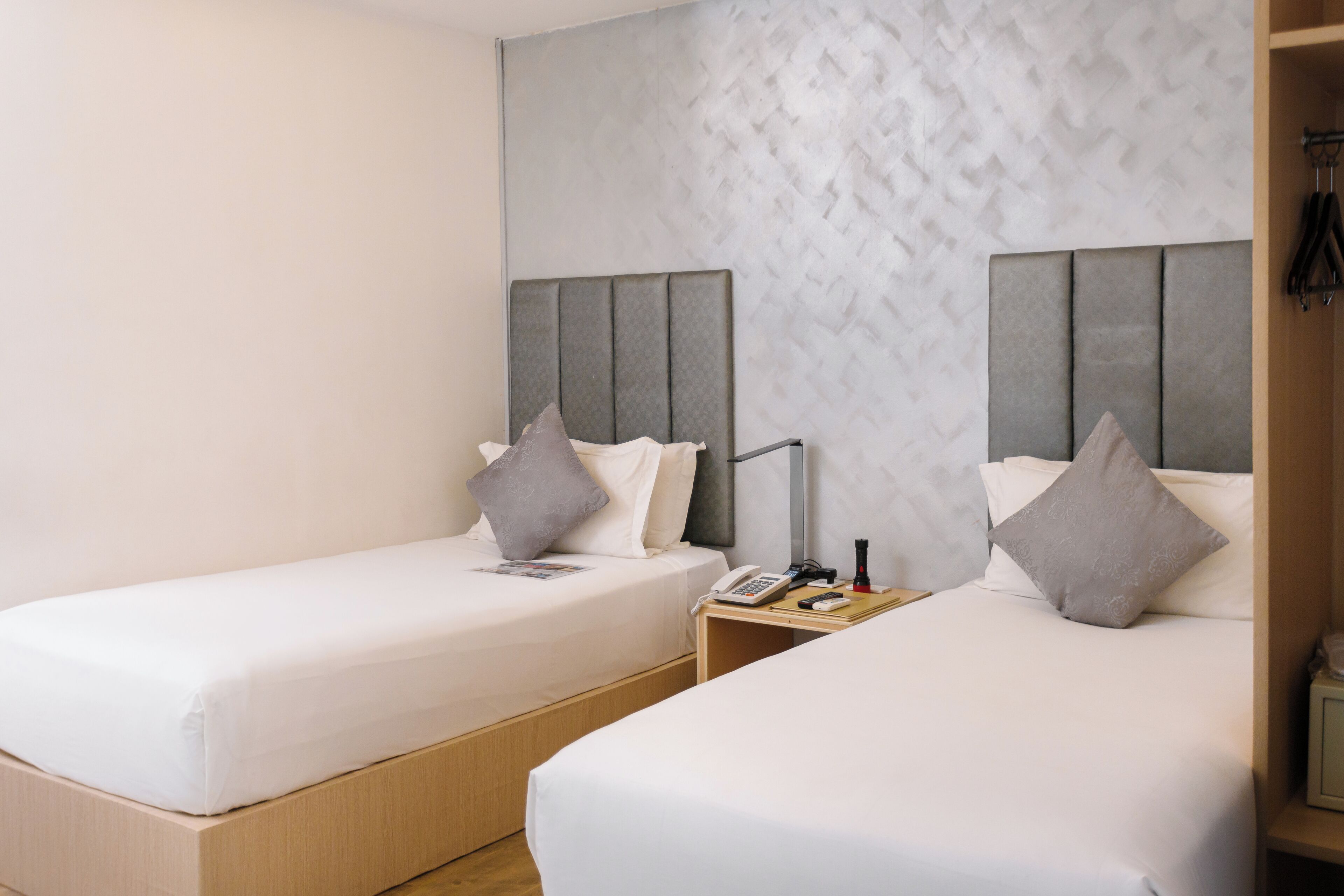 Een hotelkamer met twee enkele bedden, elk met een grijs hoofdbord, witte beddengoed en een nachtkastje met een lamp en een telefoon.
