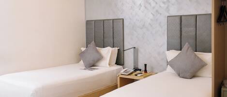 Een hotelkamer met twee enkele bedden, elk met een grijs hoofdbord, witte beddengoed en een nachtkastje met een lamp en een telefoon.