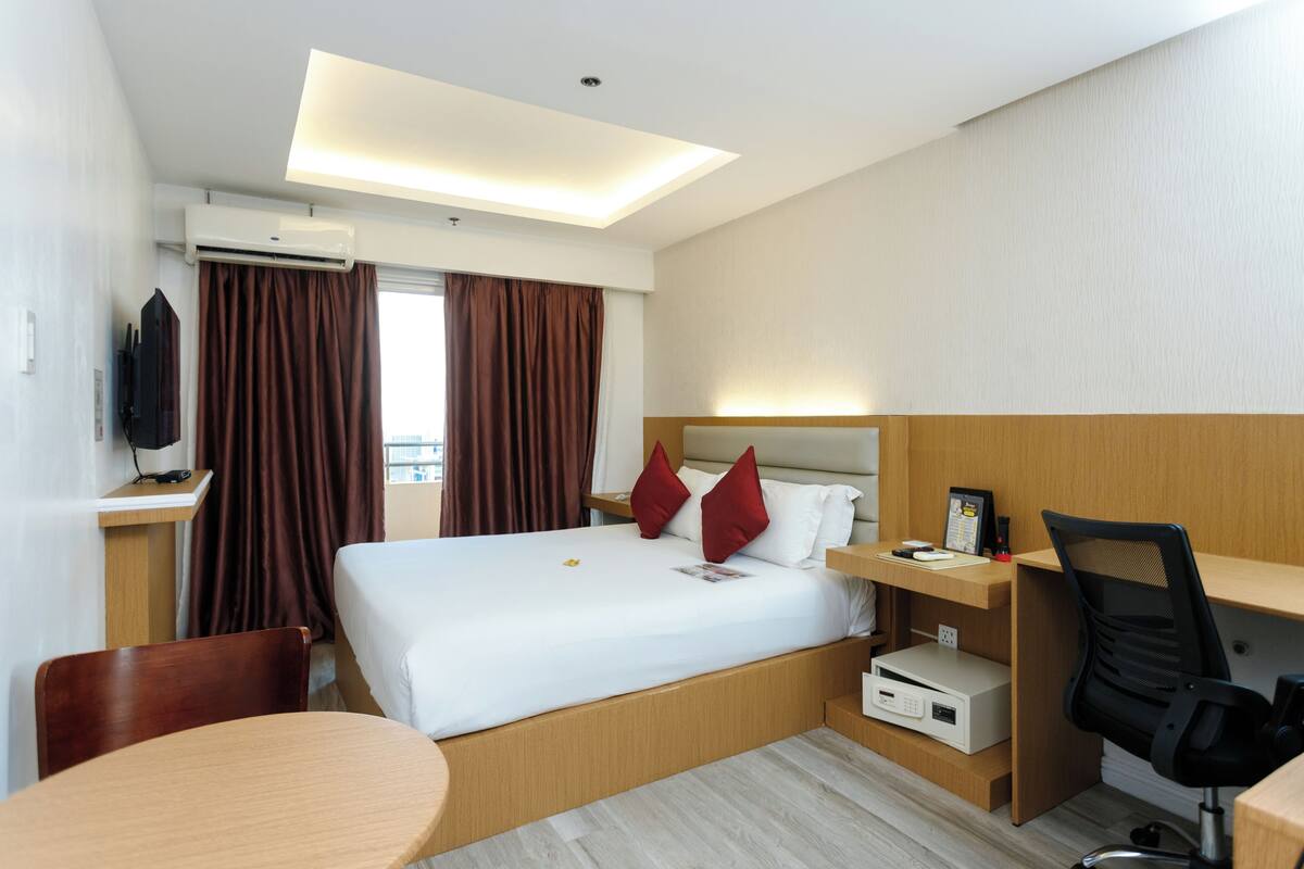 A hotel room with a bed, a desk, a chair, a TV, and an air conditioning unit.