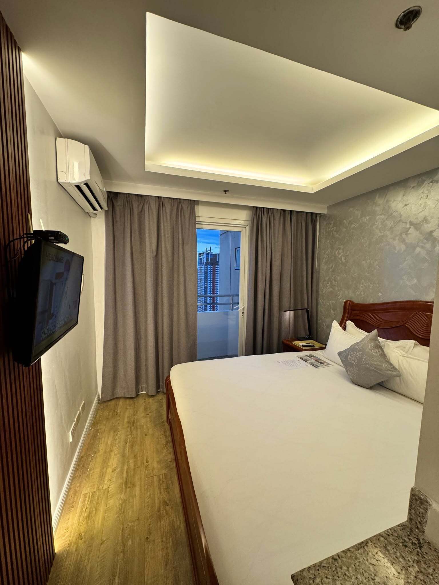 A hotel room with a bed, a television mounted on the wall, a door leading to a balcony, and an air conditioning unit.