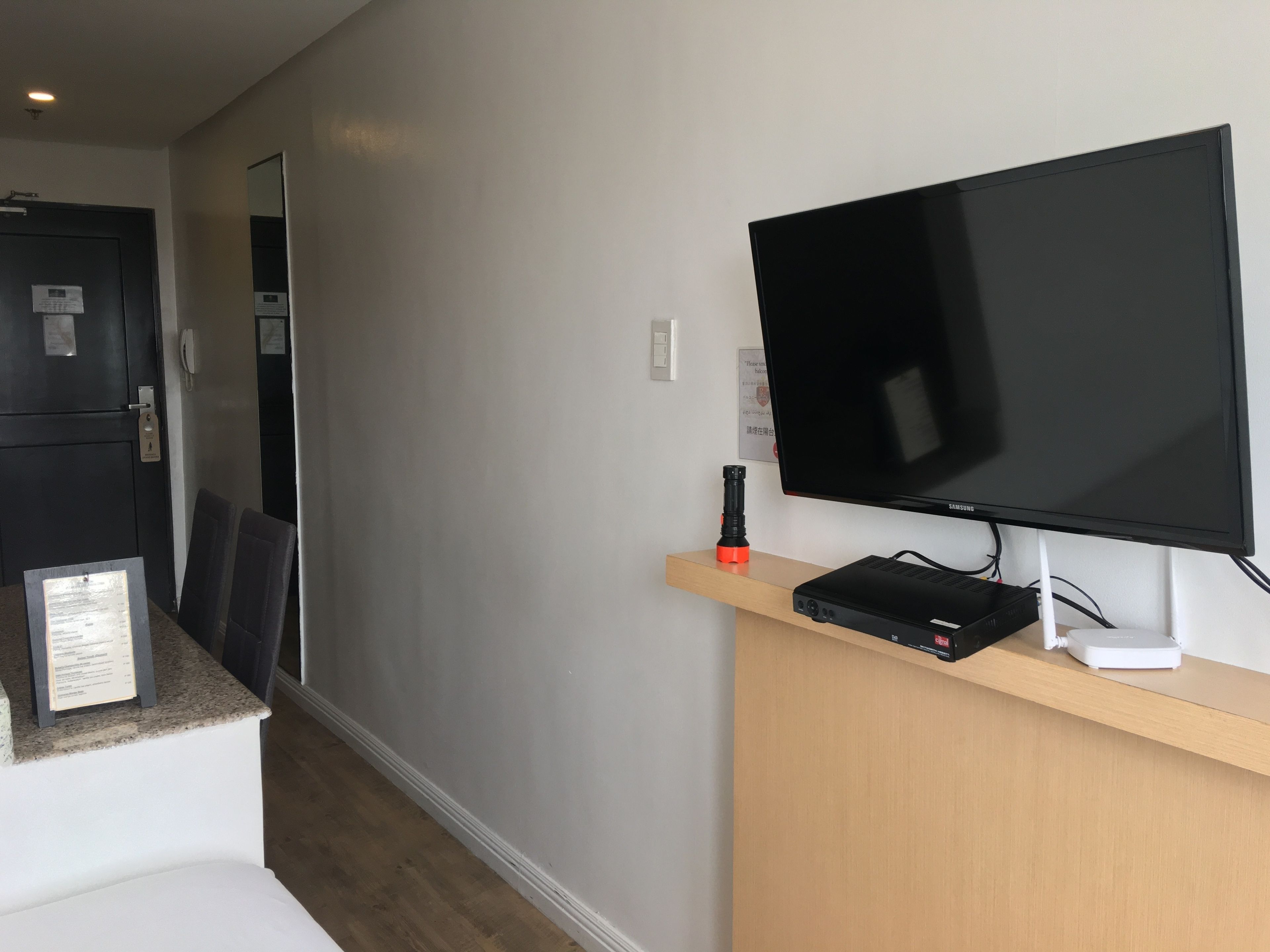 Habitación de hotel con televisor de pantalla plana sobre un soporte de madera, una mesita con un proyector y una puerta con teléfono y cerradura.