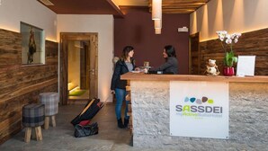 A reception desk with a sign for SASSDEI myActiveSuiteHotel. There are two people interacting at the desk, and a suitcase is visible on the floor. - Sassdei My Active Suite Hotel (Andalo)