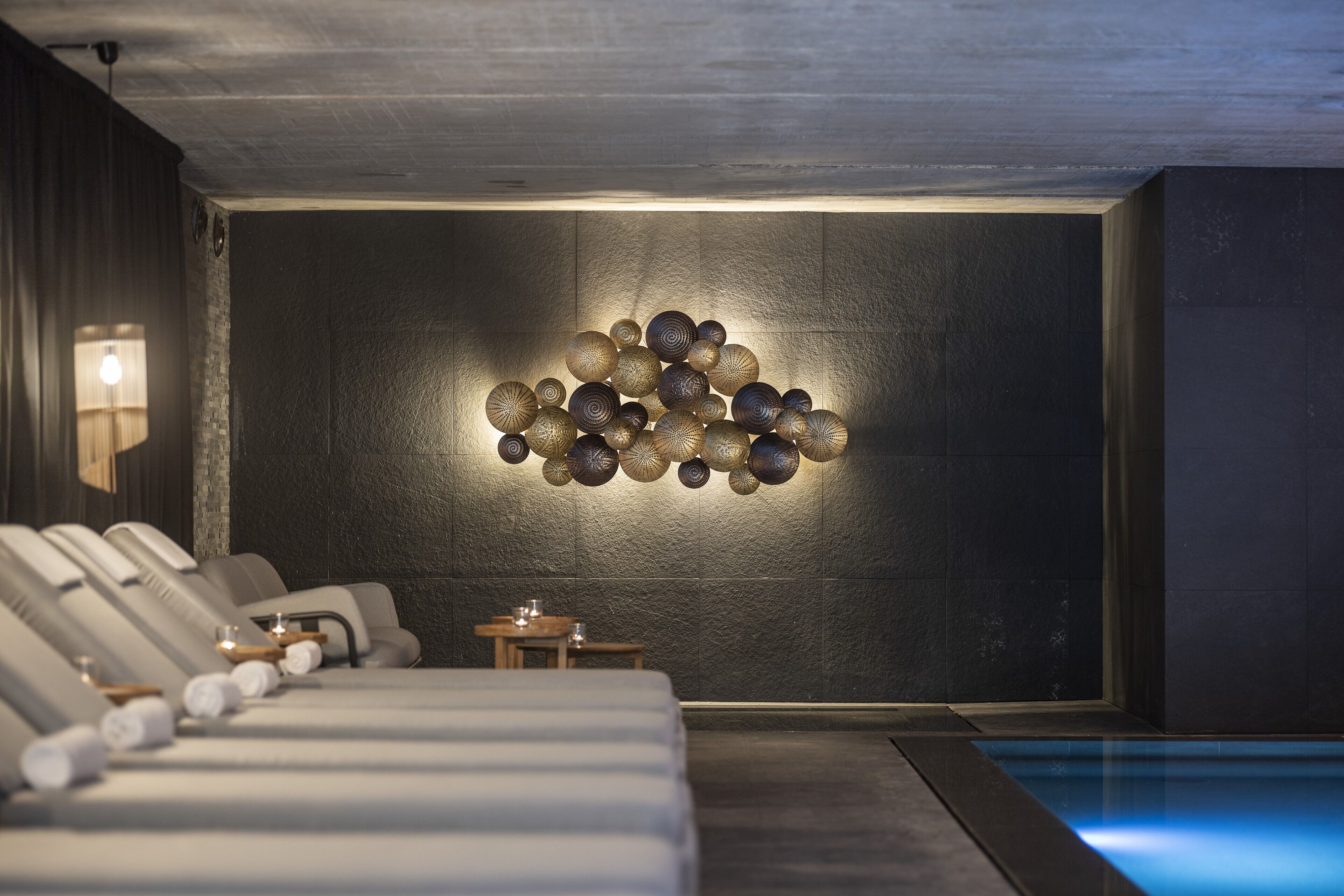 A modern spa room with a wall-mounted light fixture, a massage table, and a small table with a candle.
