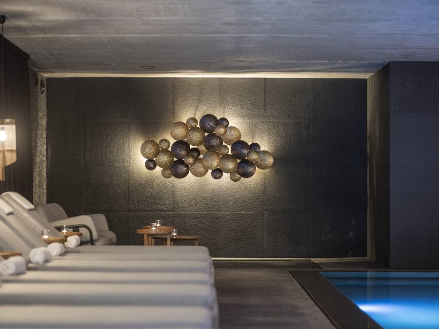 A modern spa room with a wall-mounted light fixture, a massage table, and a small table with a candle.