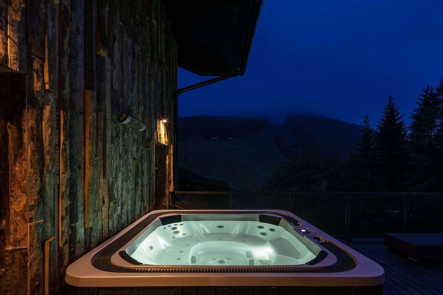 Un bain à remous à l’extérieur d’un bâtiment rustique en bois, avec vue sur une chaîne de montagnes au crépuscule.