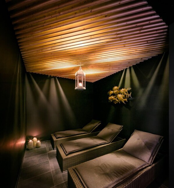 A spa room with wooden slatted ceiling, two wicker lounge chairs, a wall-mounted flower arrangement, and a hanging lantern.