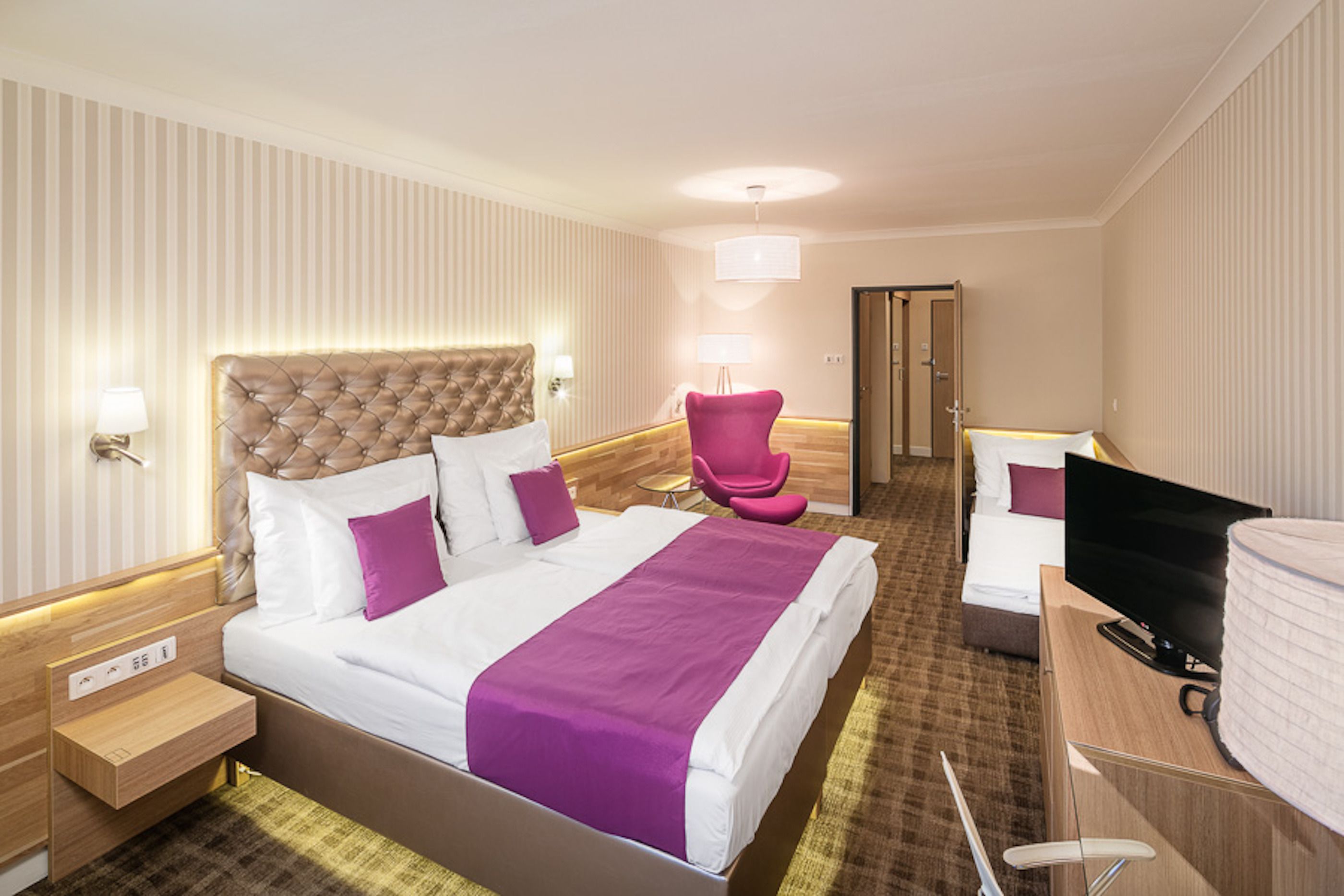 A hotel room with a large bed, a desk with a flat-screen TV, a chair, and a door.