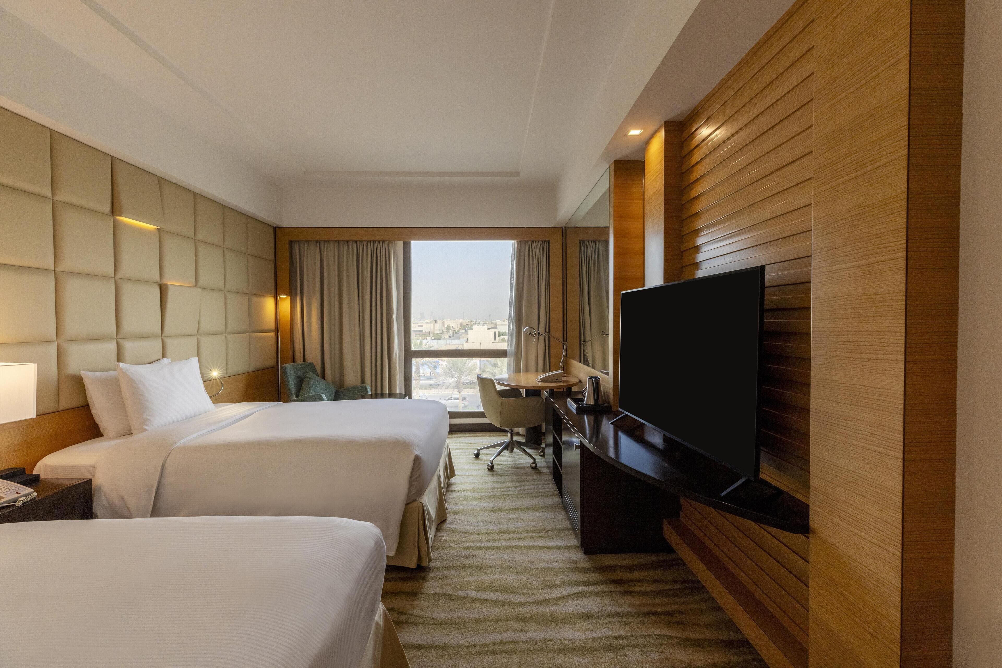 A hotel room with two beds, a desk, a television, and a view of the city.