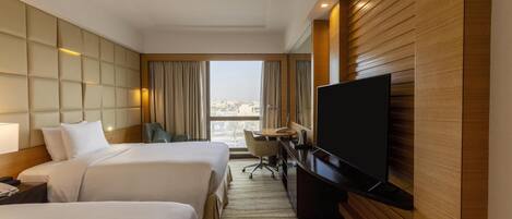 A hotel room with two beds, a desk, a television, and a view of the city.