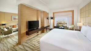 Quarto de hotel com uma cama grande, televisão, área de estar com mesa e cadeiras, e vista da cidade pela janela.