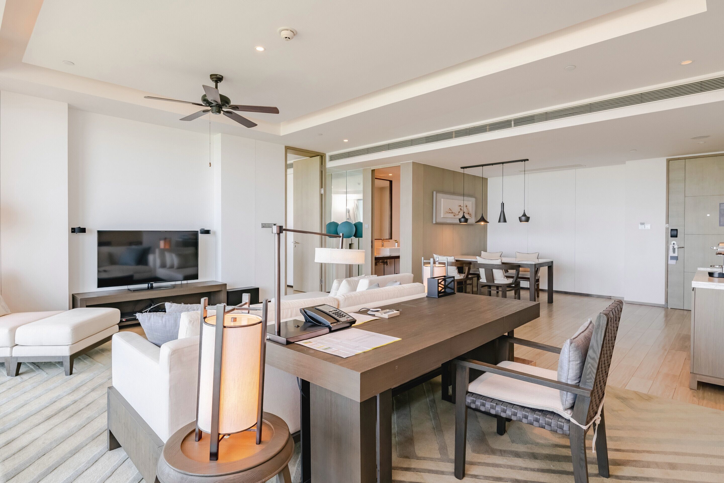 A modern hotel room with a flat-screen TV, a sofa, a dining table, and a ceiling fan.