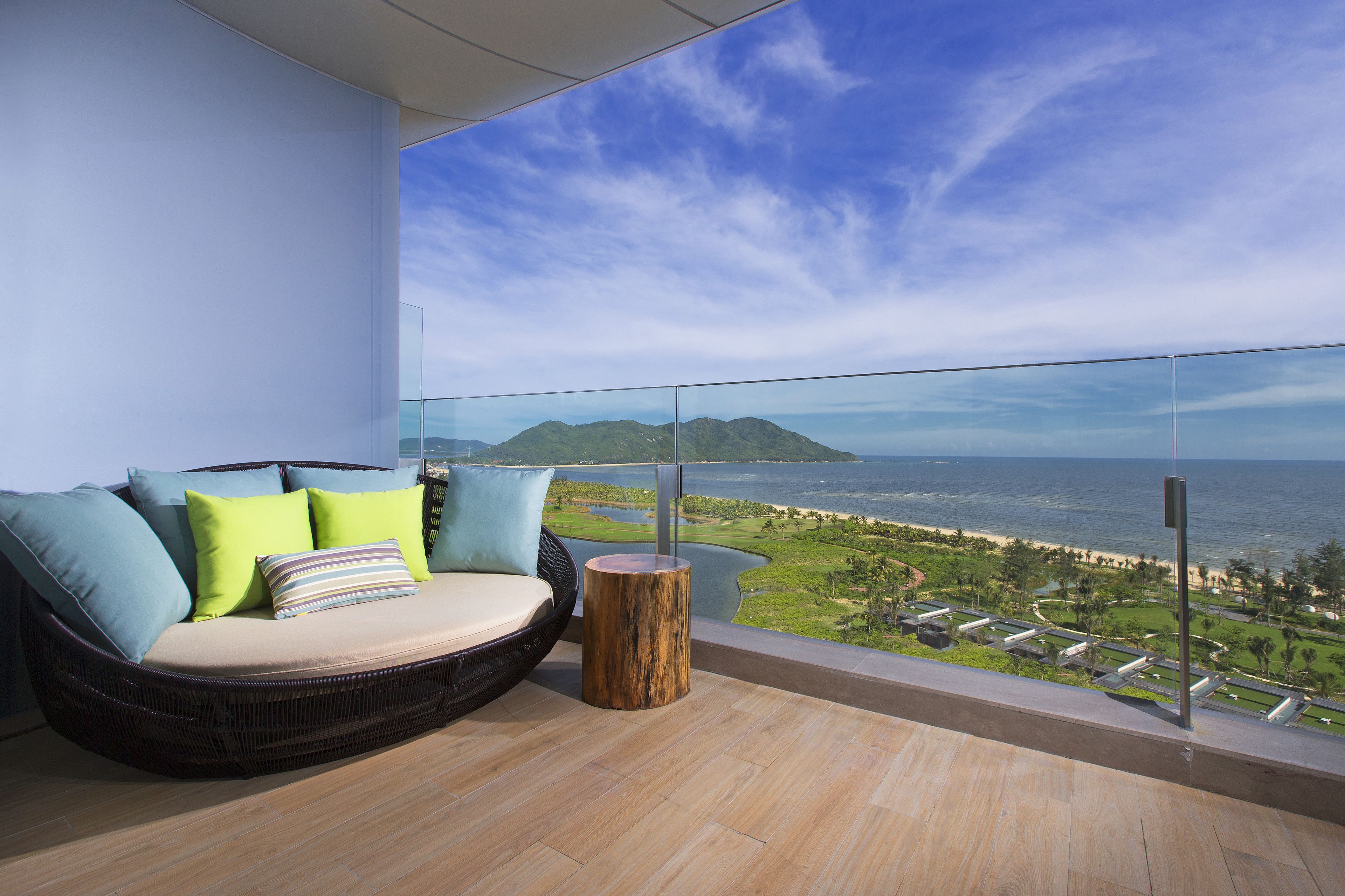 A balcony with a wicker sofa, a wooden stool, and a glass railing overlooking a coastal landscape.