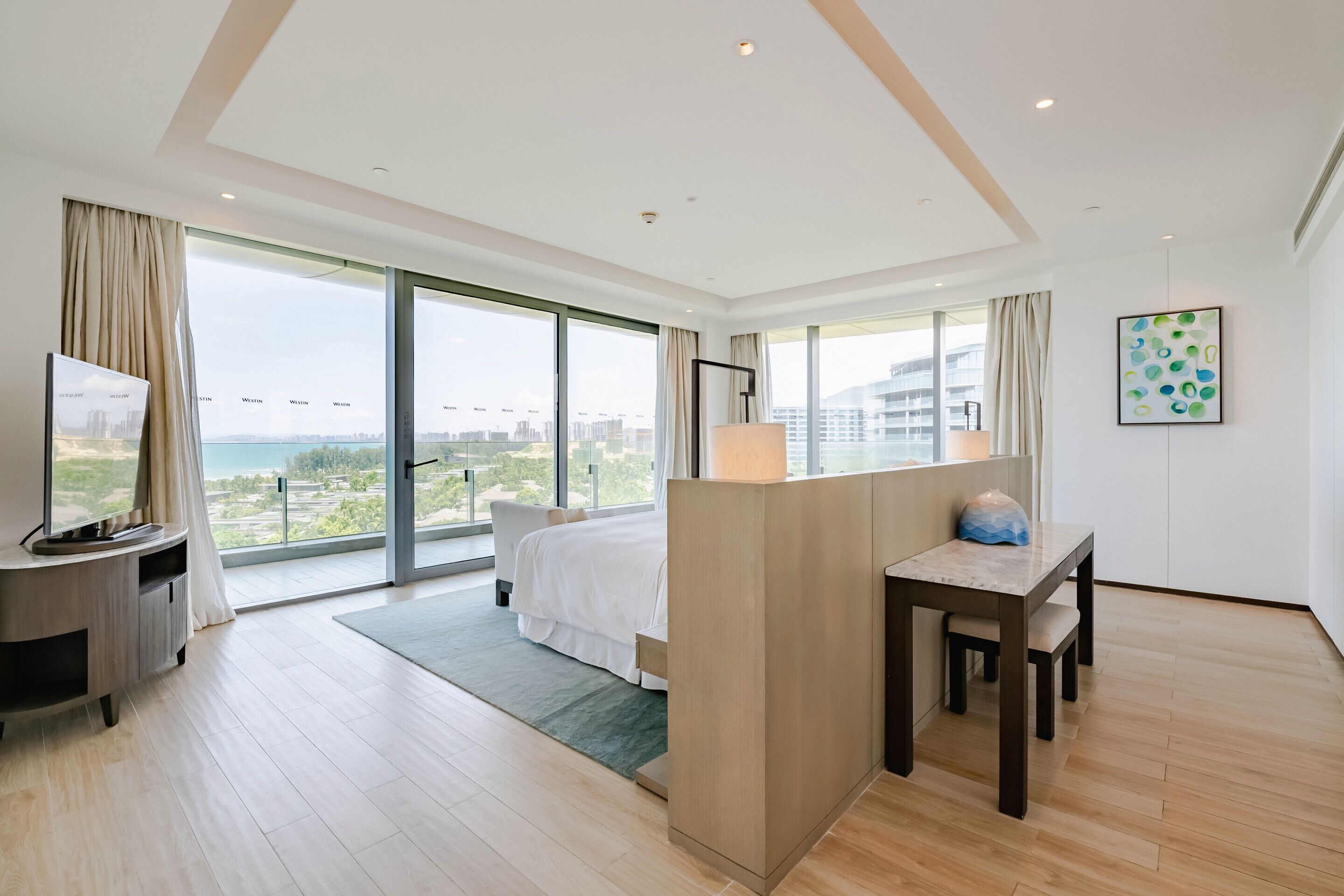 A modern hotel room with a large bed, a desk, a TV, and a view of the city and sea.