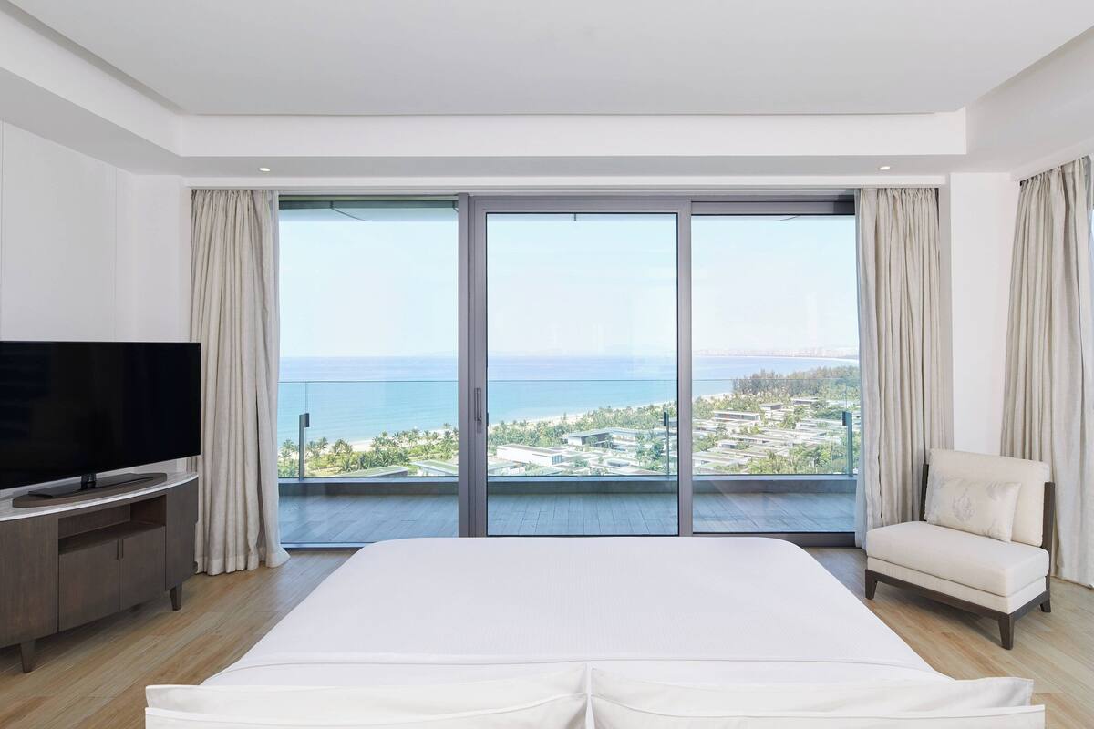 Une chambre d’hôtel avec un grand lit, un téléviseur posé sur un meuble en bois, et une vue sur la mer grâce à de grandes fenêtres du sol au plafond.